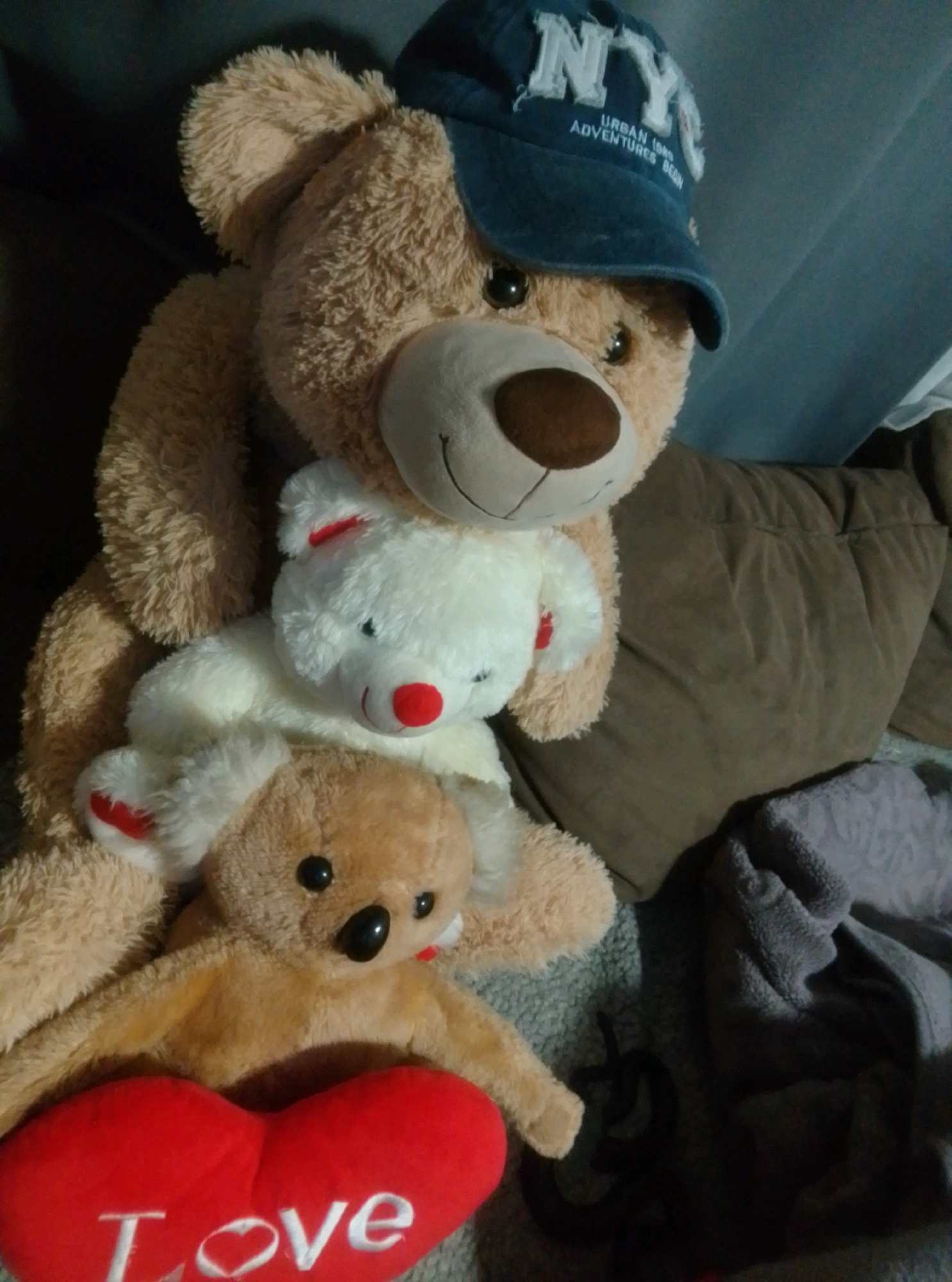 A group of stuffed teddy bears are arranged on a sofa. The largest teddy bear, positioned at the top and center of the image, is light brown and wears a blue baseball cap with white lettering that reads "NYC URBAN 1999 ADVENTURES BEGIN". It has a black nose and visible stitching for its mouth. Beneath it, a smaller white teddy bear with a red nose and red accents on its ears and paws is partially visible. At the bottom center of the image, a light brown teddy bear lies down, with its head resting on a red heart-shaped pillow that has the word "Love" embroidered in white. The sofa appears to be covered in a brown fabric. To the right, a dark gray textured blanket is visible. The lighting in the scene is dim. The context of Wedel, Germany, is not discernible from the visual information provided in the image.