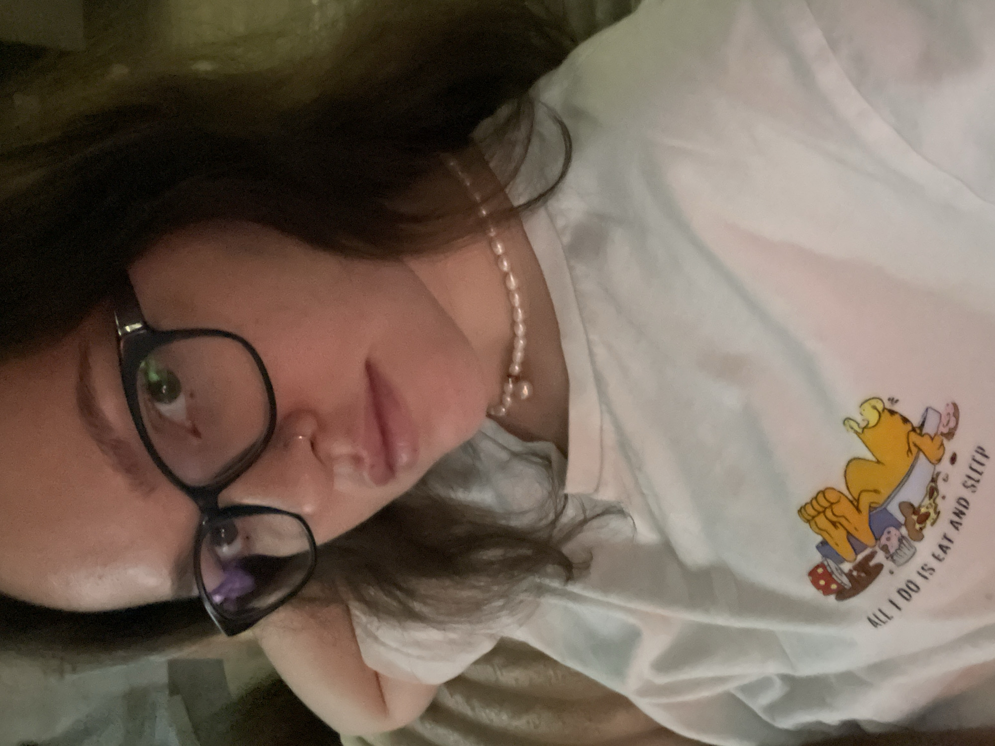 A close-up, angled portrait features a young person, likely female, lying down in an indoor setting with dim, soft lighting, suggesting evening or night. The individual has long, dark hair visible around their head and shoulders. They are wearing black-framed glasses, which show reflections, and have a small silver hoop piercing on their nose. A pearl necklace with a small golden charm is worn around their neck.
The person is clad in a white t-shirt that displays a colorful cartoon character, an orange cat resembling Garfield, lying on its back surrounded by various food items including cupcakes, cereal, and a slice of pizza. Adjacent to the character, the text "ALL I DO IS EAT AND SLEEP" is printed vertically.
The subject looks directly towards the camera with a neutral and relaxed expression. The background immediately behind the head is dark and out of focus, while a lighter, textured fabric, possibly a blanket or pillow, is visible underneath the person. The image captures the subject's upper body and face. This scene is set in Izhevsk, Russia.
