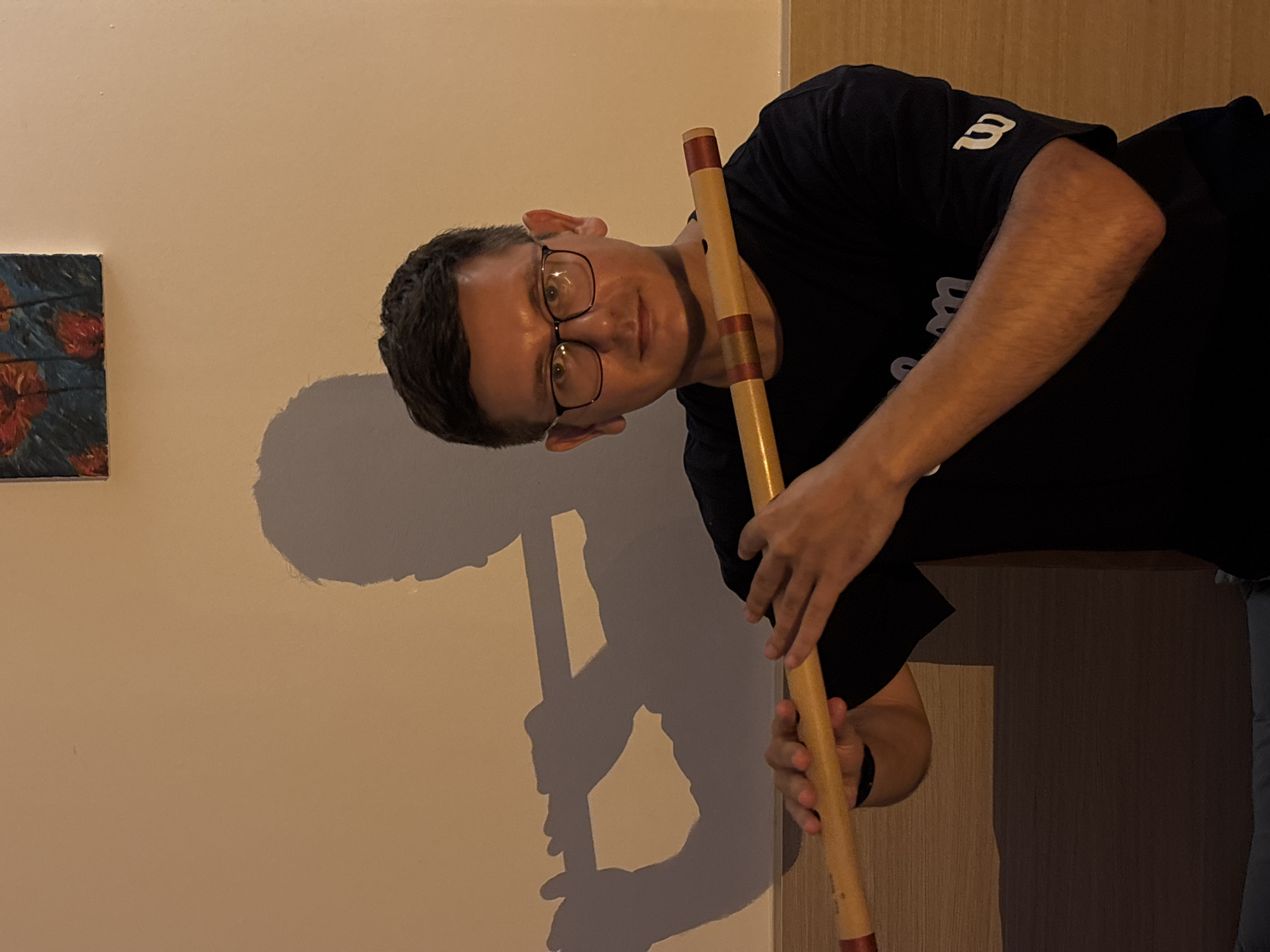 A man with glasses and dark hair plays a wooden flute in front of a light-colored wall. His shadow is cast on the wall behind him, showing him holding the flute to his lips. He is wearing a black t-shirt with a white logo on the sleeve. The background is a plain wall with a small framed picture of flowers hanging above.