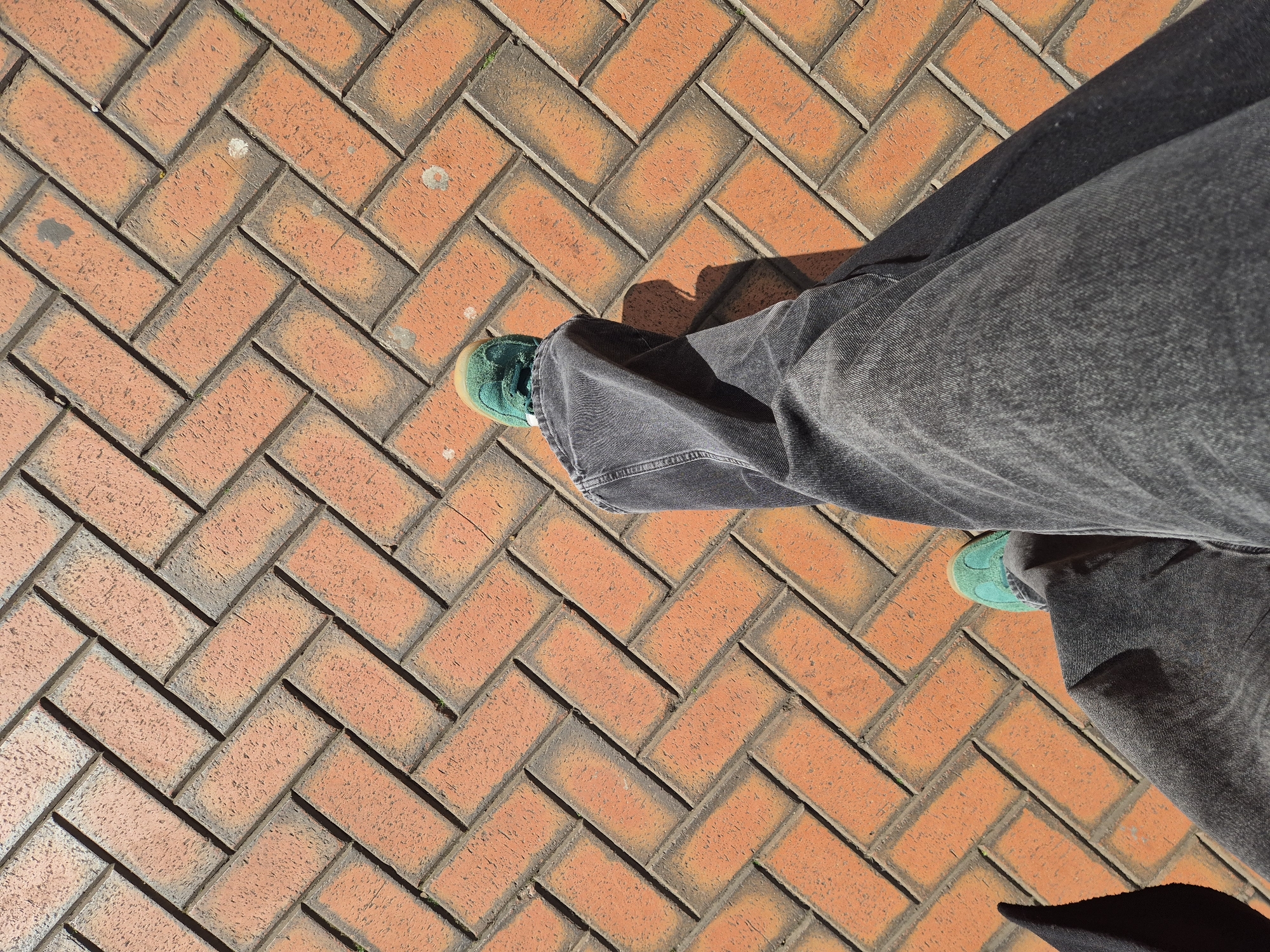 The image shows a view from above of a person standing on a brick-paved surface. The bricks are arranged in a herringbone pattern and are predominantly orange-red with darker outlines. The person is wearing wide-legged, gray denim jeans and bright green sneakers with a light-colored sole. The jeans are partially obscuring the view of the bricks. A shadow is visible near the lower right corner of the frame. The location is a brick paved footpath somewhere in Chelmsford, United Kingdom.
