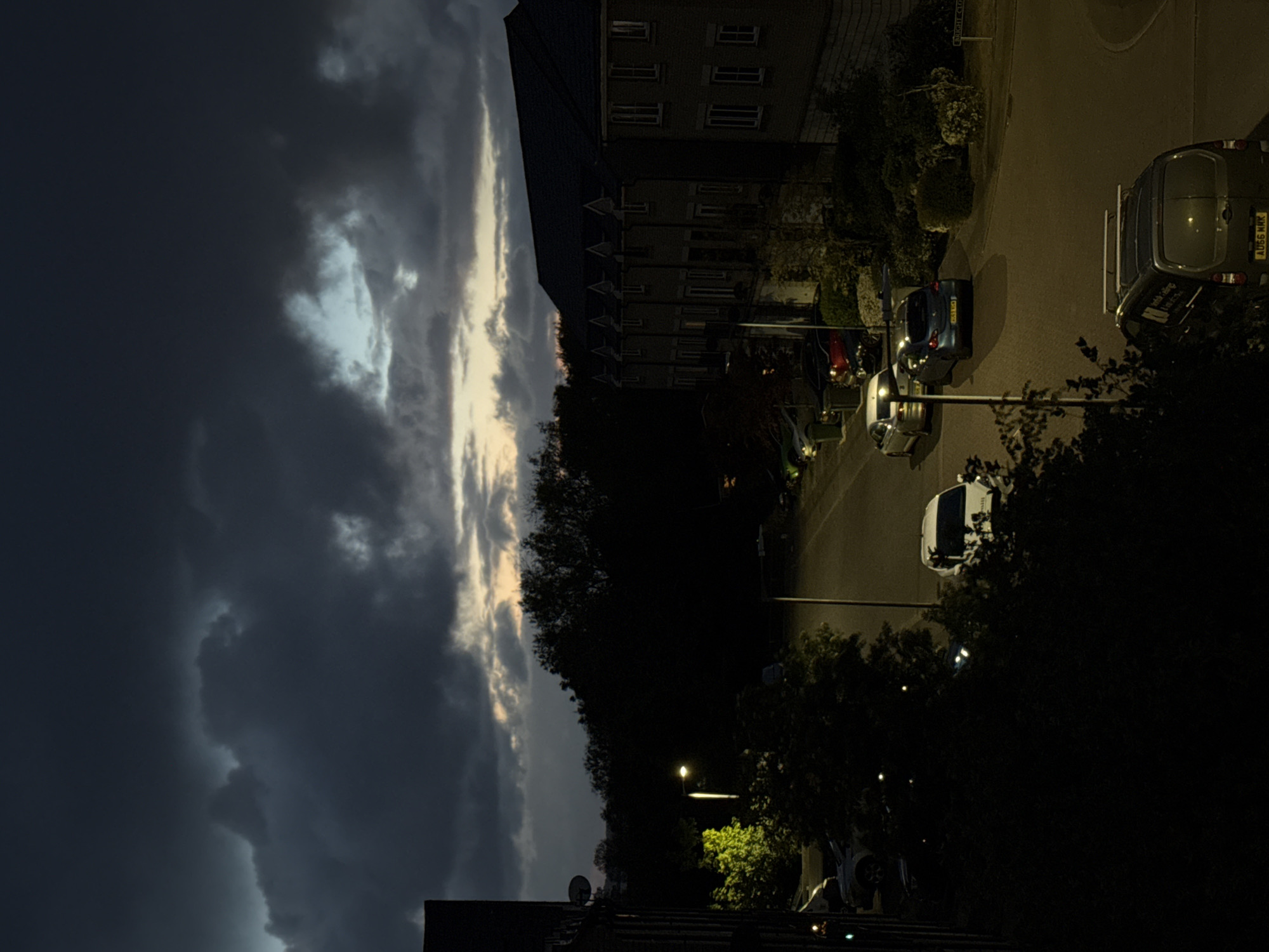 A nighttime scene in Great Yarmouth, United Kingdom, shows a street with parked cars under streetlights. A building stands along the street with multiple lit windows. The sky is filled with dark, heavy clouds and a sliver of light. Trees line the street, some obscuring the view. One van and multiple cars can be distinguished, including a white car and a blue car.