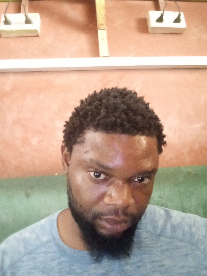 An adult male with dark skin, short curly hair, and a full dark beard is depicted in a close-up portrait in Magboro, Nigeria. He wears a light grey or blue textured short-sleeved t-shirt and has a small gold hoop earring in his left ear. His gaze is directed intensely and directly at the camera, with a visible sheen on his forehead. The background is a reddish-brown textured wall. Mounted on the upper section of the wall are two white electrical outlets, each with two dark plugs inserted. A horizontal white strip runs below these outlets, and a vertical light-colored wooden beam is positioned to the right of the frame's center. A faint green element is partially visible in the lower-left background.