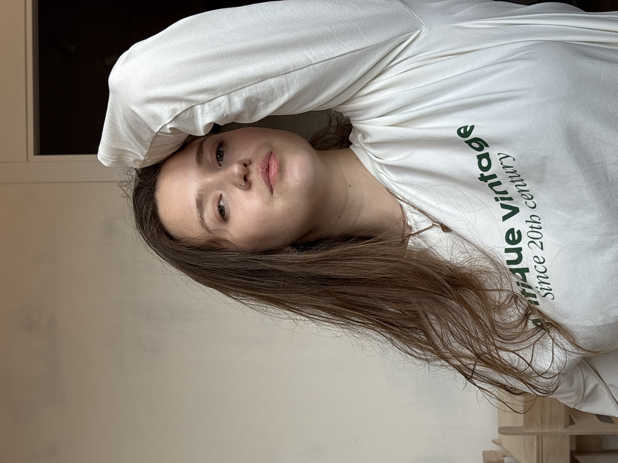 A woman with long brown hair, a nose ring, and light skin is lying down, facing the camera. She is wearing a white long-sleeved shirt with green text that reads "Antique Vintage since 20th century". Part of a window is visible in the top left corner. A white wall is behind her. There are wooden objects or furniture visible on the bottom right side of the image. The location in the city of Izhevsk, Russia, is not determinable from the image.