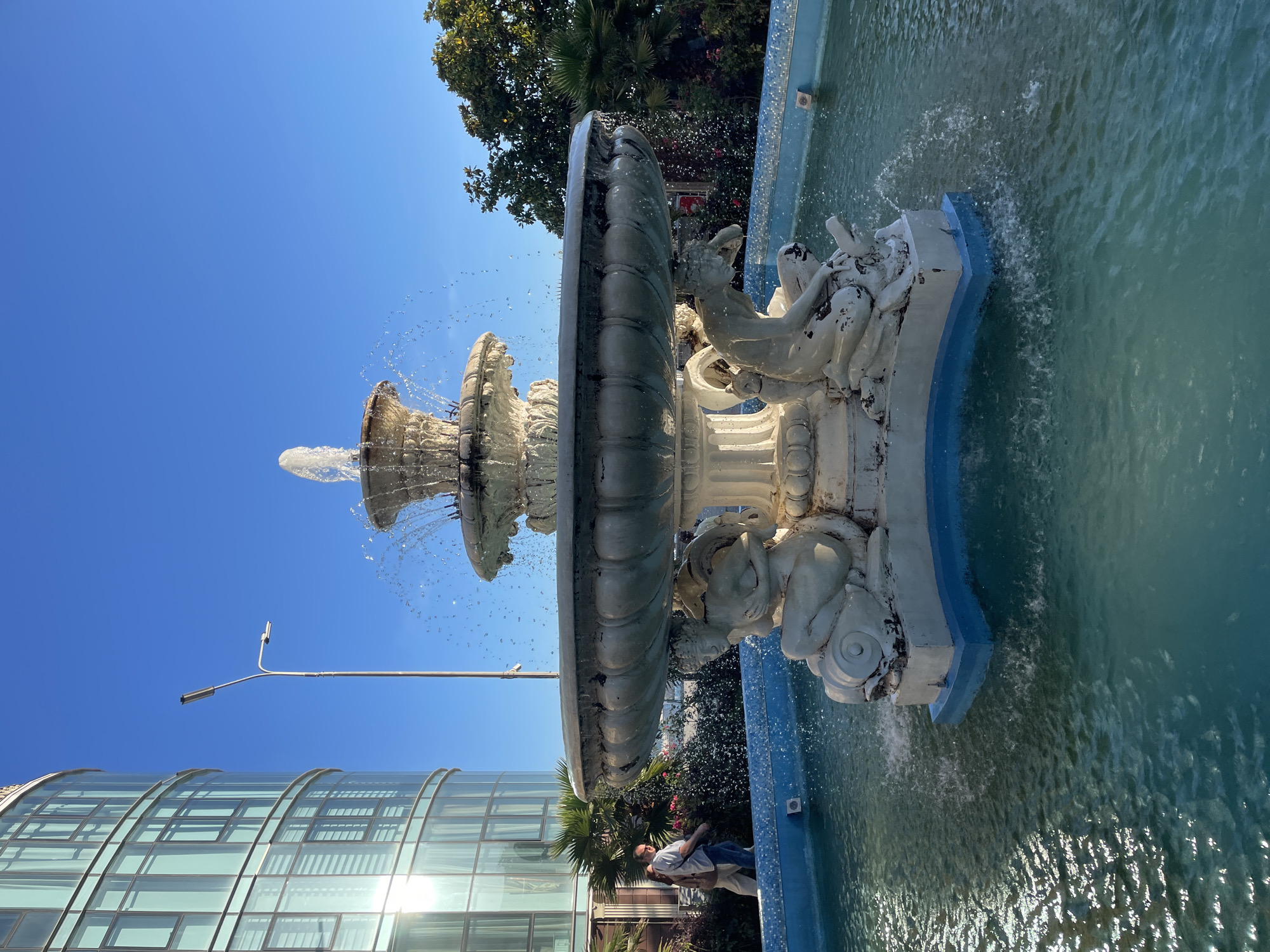 The image captures a multi-tiered, ornate fountain in a city square, likely in Sochi, Russia, given the architectural style of the building and the presence of palm trees. The fountain, made of white stone, features sculptures of figures and mythical creatures, with water cascading from its upper levels into a large, turquoise basin. The water in the basin is clear and reflects sunlight, with ripples and splashes indicating the active flow of the fountain.
To the left of the fountain, a modern, curved building with a glass facade dominates the background. The building's windows reflect the bright blue sky and the sun's glare. A street lamp with a contemporary design stands between the building and the fountain.
Two people are walking in the background, near a lush green area with palm trees and flowering bushes. The sky is clear and a vibrant blue, suggesting a sunny day. The overall scene conveys a sense of urban elegance and public space.