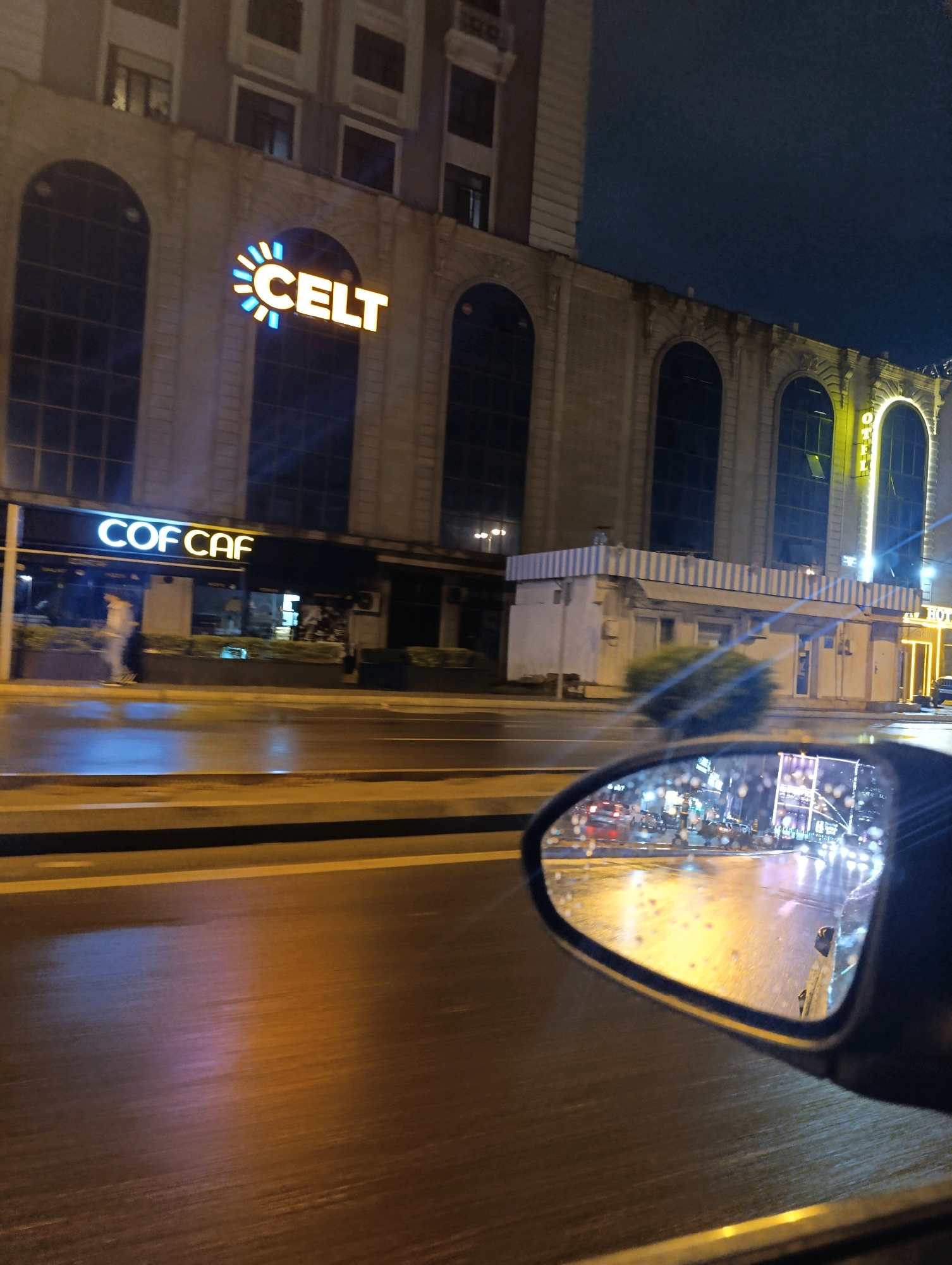 This image depicts a nighttime street view in Baku, Azerbaijan, taken from a moving vehicle. The foreground features a wet road reflecting the city lights, with a car's side mirror in the lower right corner showing a distorted, illuminated reflection of the street and buildings. Directly ahead, a multi-story building with arched windows is illuminated by signage. On the building's facade, a large sign reads "CELT" with a stylized sun logo. To the left, another illuminated sign displays "COF CAF," indicating a cafe. The overall environment is urban, with streetlights, vehicle lights, and building lights contributing to the nighttime ambiance. The wet road suggests recent rain.