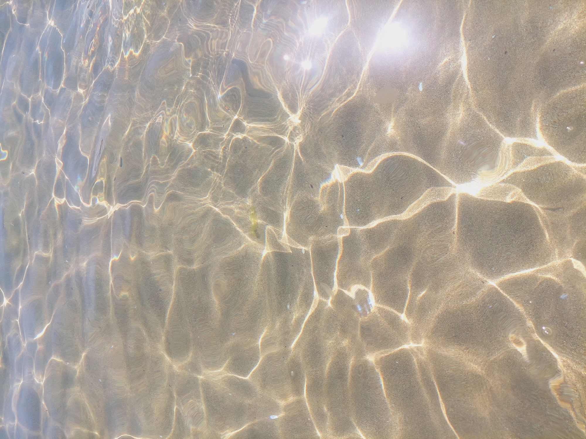 The image depicts a close-up, overhead view of shallow, clear water with sandy bottom. Sunlight refracts through the ripples on the water's surface, creating a pattern of bright, shimmering light and darker shadows on the sand. The sand appears to be a light brown or tan color, with some small pebbles and debris visible. There are no people, animals, or man-made objects in the frame. The environment is natural, suggesting a beach or shallow sea. The location is the coast of Sozopol, Bulgaria.