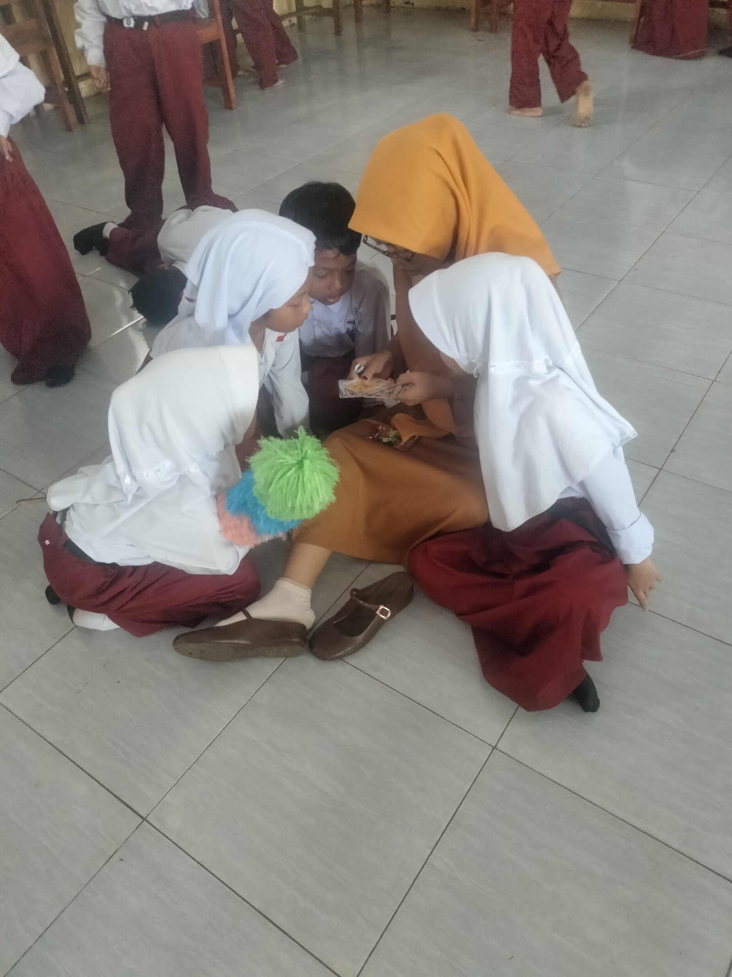 In a classroom in Gunung Tugel, Indonesia, several children in school uniforms are gathered on the floor, interacting with items. Three girls in white hijabs and maroon skirts are seated, with one leaning forward, examining something in her hands. Another girl, wearing a yellow hijab, is also seated and looking down. A boy in a light blue shirt is seated behind them, observing. The floor is tiled in a light gray color. The scene suggests a group activity or a shared moment of observation among the students.
