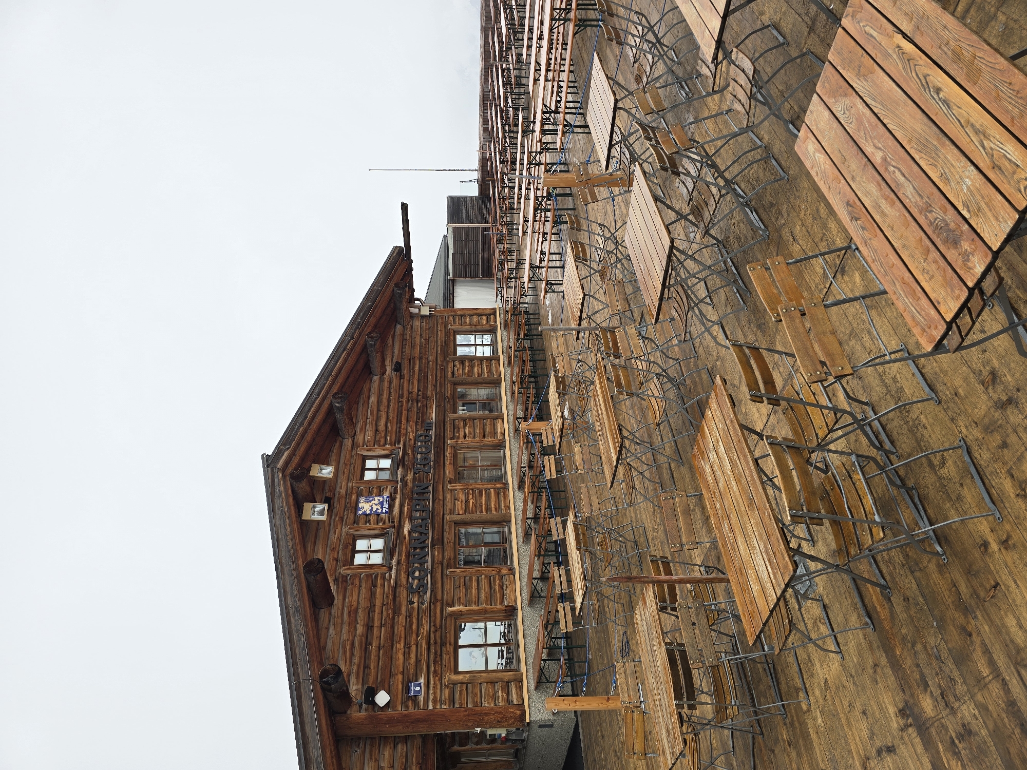 The image shows the exterior of the Sonnalpin restaurant on the Zugspitze, Germany's highest peak, located in Garmisch-Partenkirchen. The restaurant is a multi-story wooden structure with a prominent pitched roof. The building's facade features windows and a sign with the restaurant's name. An outdoor seating area with wooden tables and chairs is adjacent to the building. The sky is overcast.