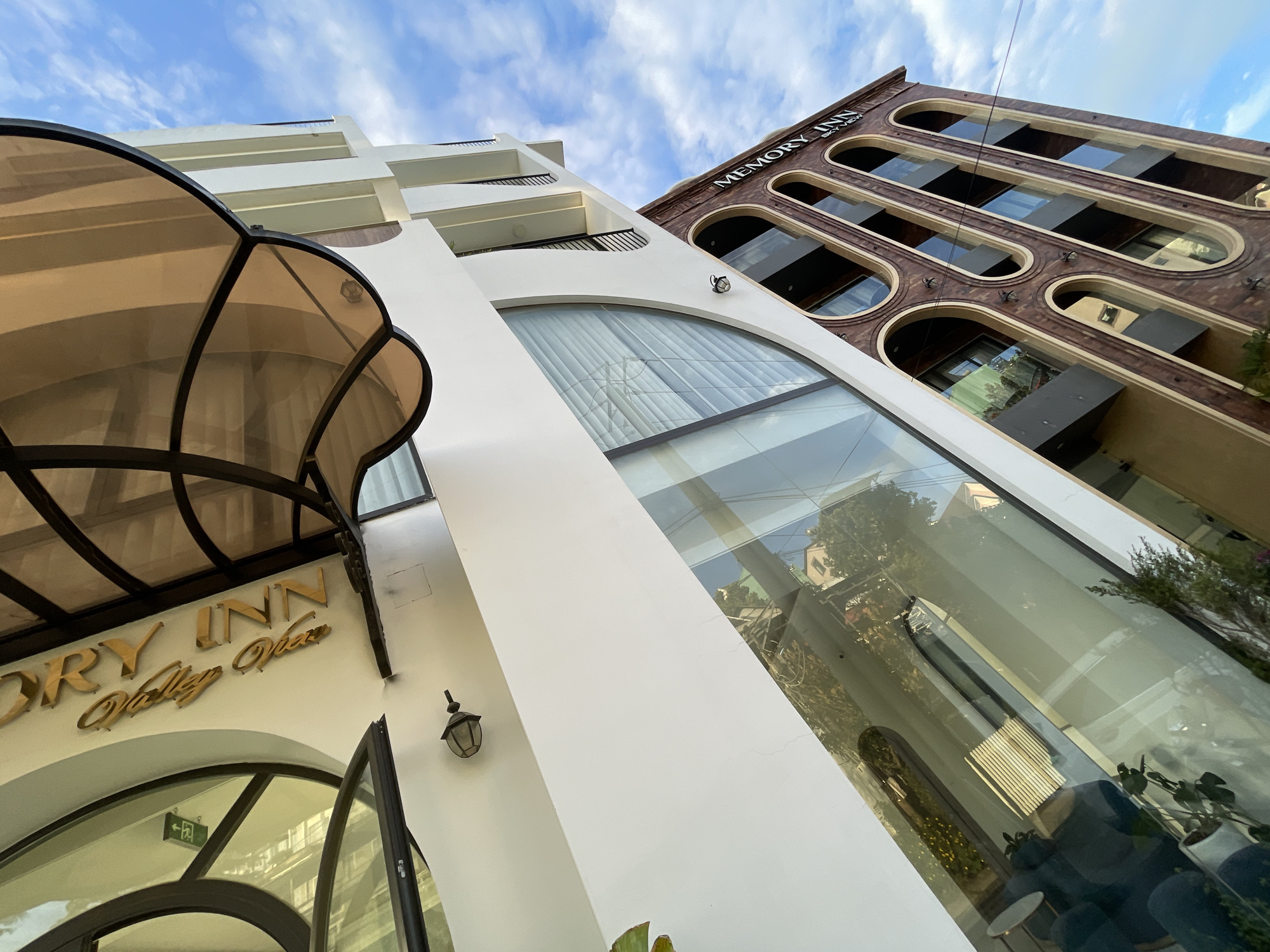 The image shows the exterior of the Memory Inn Sky View hotel in Da Lat, Vietnam. The hotel has a modern architectural design with a white facade and a darker, brick-like lower section featuring arched windows. The sky is partly cloudy and blue. The "MEMORY INN SKY VIEW" sign is visible on the darker section of the building. The image is taken from a low angle, looking up at the hotel.