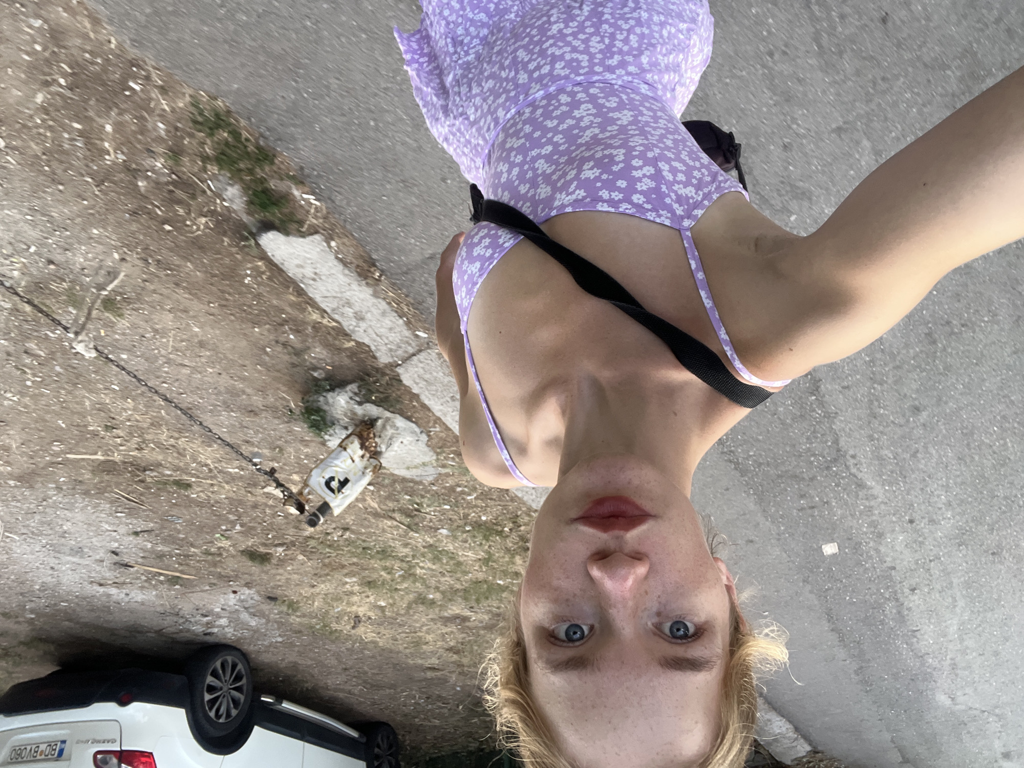 The image shows a young Caucasian woman with blonde hair and blue eyes, taking a selfie outdoors. She is wearing a light purple floral print dress and a black strap is visible across her chest. She is standing on a paved surface that appears to be a road or sidewalk. In the background, a white SUV with a license plate that reads "BD BV060" is parked. There is also a rusty metal post with a chain attached, and dry vegetation and dirt on the ground. The scene is consistent with a location in the city of Budva, Montenegro, a coastal town known for its scenic roads and vehicles with local license plates.