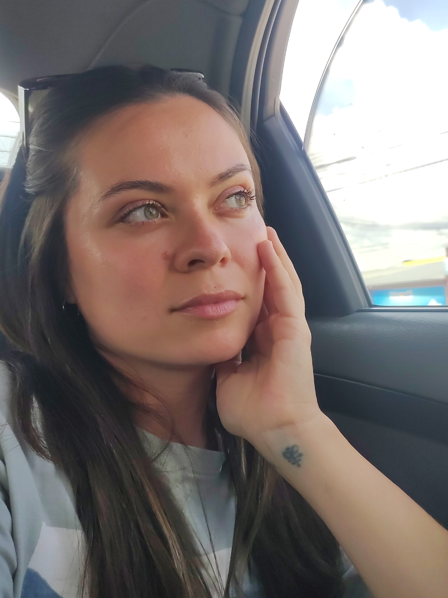 A young woman with long, dark hair and light eyes is looking out a car window. She is wearing a light-colored shirt and has her right hand on her cheek. There is a small tattoo on her left wrist. The background shows a blurry cityscape.