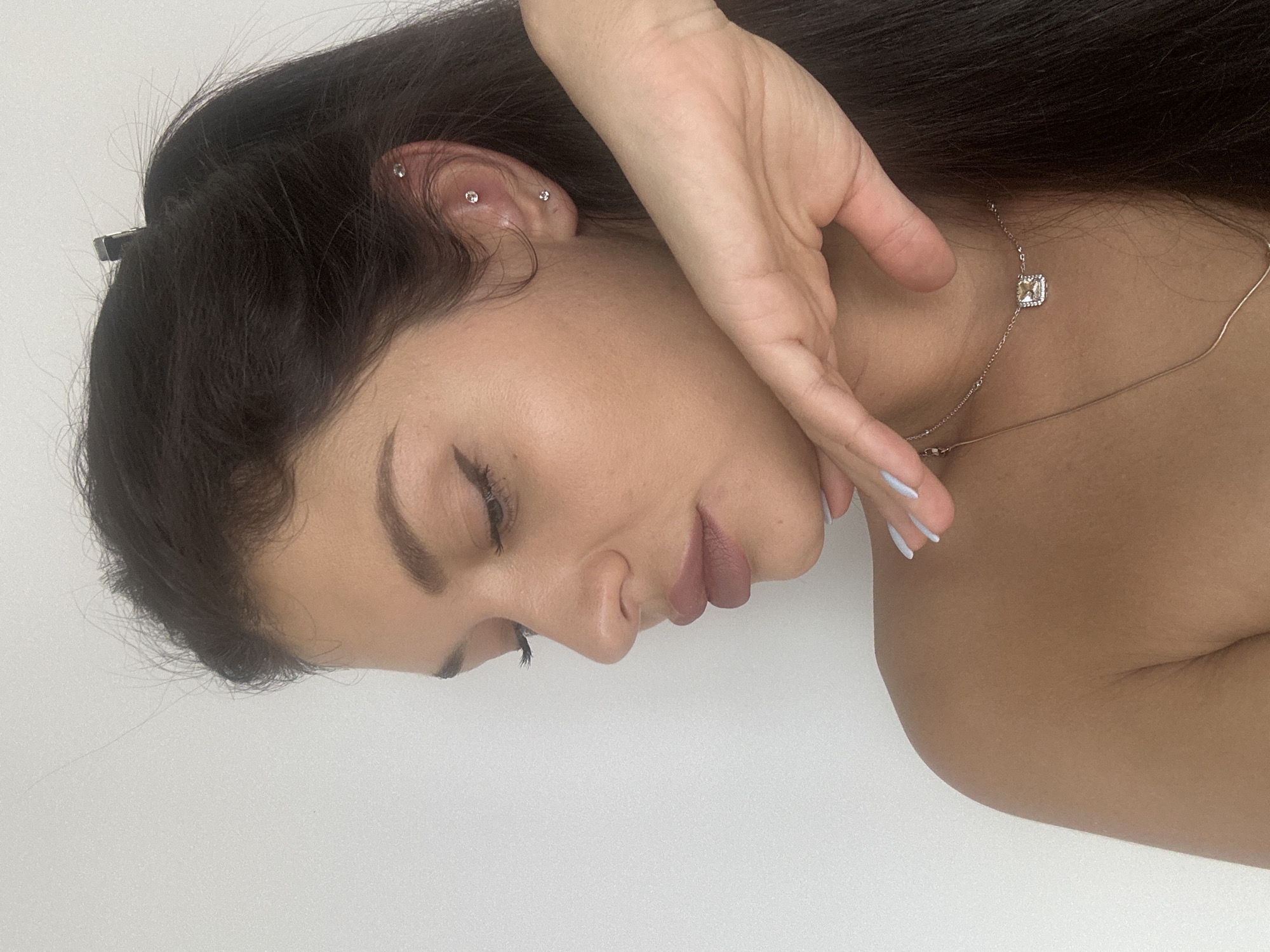 The media file features a close-up profile view of a young woman with dark hair, rendered in a vertically oriented portrait. Her eyes are closed, imparting a serene or contemplative expression. Her dark hair is partially held back by a small, dark hair clip visible near her ear.
Her ear is adorned with three small, sparkling stud earrings; two are positioned along the upper cartilage (helix) and one is slightly lower. A delicate, silver-toned necklace with a small, square gemstone pendant rests around her neck. Her right hand is gently placed near her face and neck, with her fingernails painted in a light blue shade. She wears a natural-toned lipstick on her lips. Her skin has a warm, medium complexion.
The background is a plain, light-colored surface, likely a white wall, indicating an indoor setting. There is no visible text within the image. The scene conveys a sense of calm and understated elegance. The provided context indicates the location is Moscow, Russia.