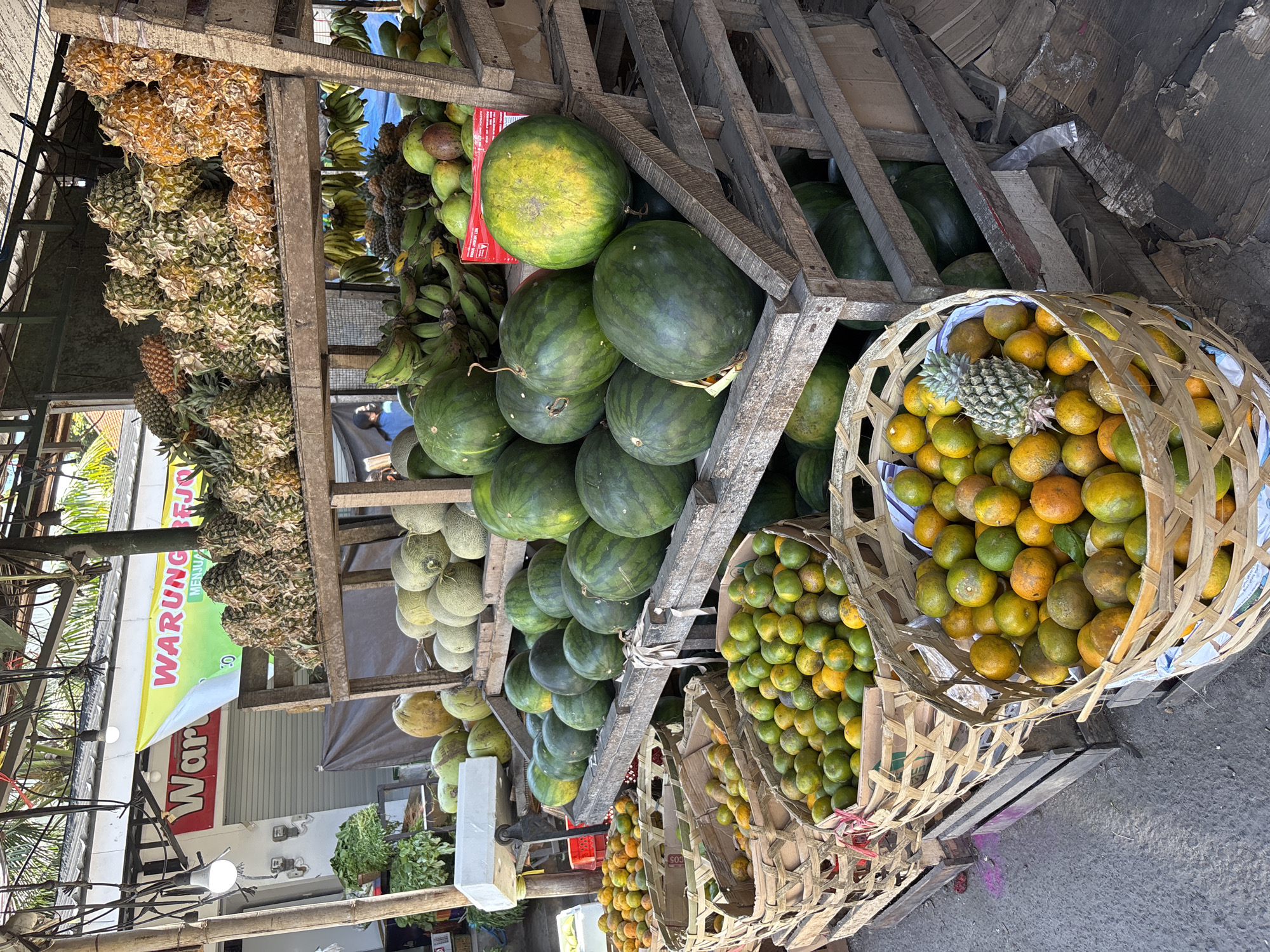 The image depicts a fruit stand in Ungasan, Indonesia. The stand is constructed of wood and is stocked with pineapples, bananas, watermelons, cantaloupes, and oranges. The fruits are arranged in baskets and wooden crates. Two woven baskets are filled with oranges, with one pineapple nestled in one of them. The word "WARUNG" is visible on a sign, suggesting a small local business. Power lines are visible above, and other shops can be seen in the background. The scene is set on a paved street.