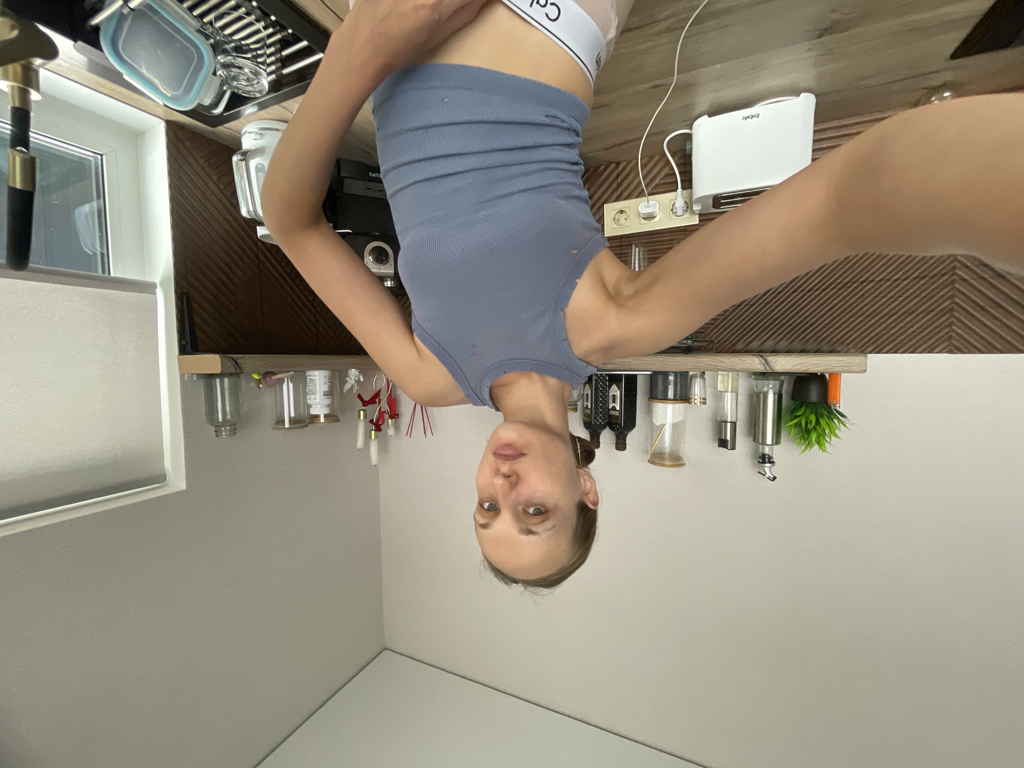 The image shows a woman in a kitchen. She is wearing a gray tank top and white underwear. The kitchen has wooden cabinets and a white countertop. There is a toaster, an electric kettle, and a dish rack on the counter. The woman is holding a camera or phone, taking a selfie. The background shows shelves with various items, including bottles, jars, and decorative objects. The lighting is natural, coming from a window on the right side of the image.