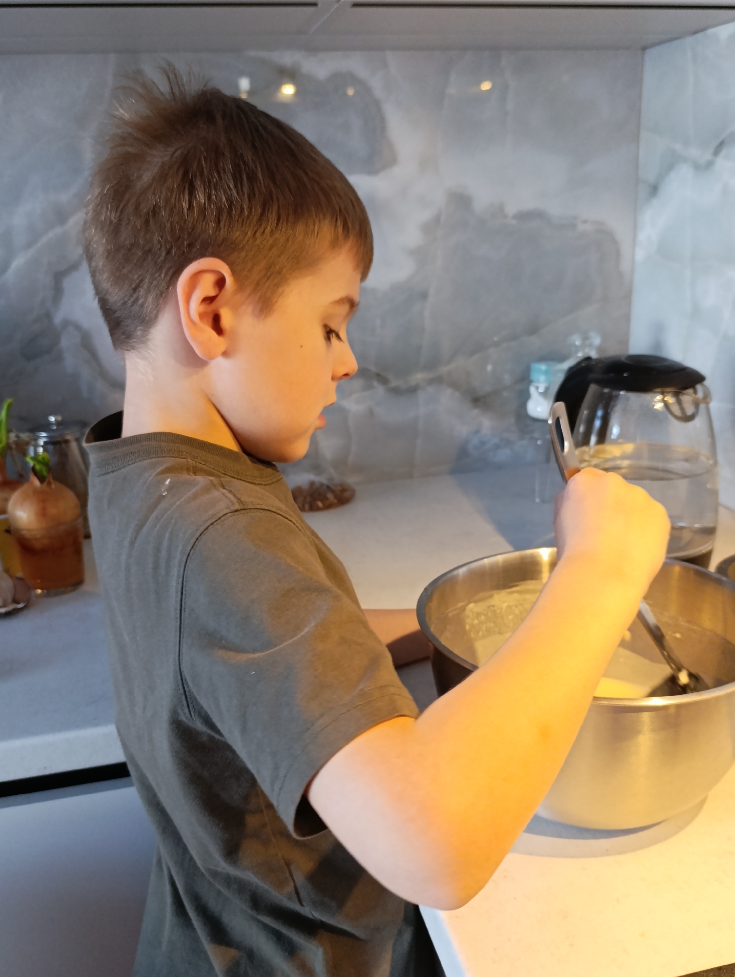 A young boy stands in a kitchen, stirring a mixture in a large, stainless steel bowl with a spoon or ladle. He wears a dark grey t-shirt. The background reveals a kitchen setting with a grey marble-patterned backsplash. A glass kettle filled with water is visible near the bowl. There is an onion on the counter, and some other objects in a glass jar. The image was most likely taken indoors. There are no details allowing to identify the location within Chaikovsky, Russia.