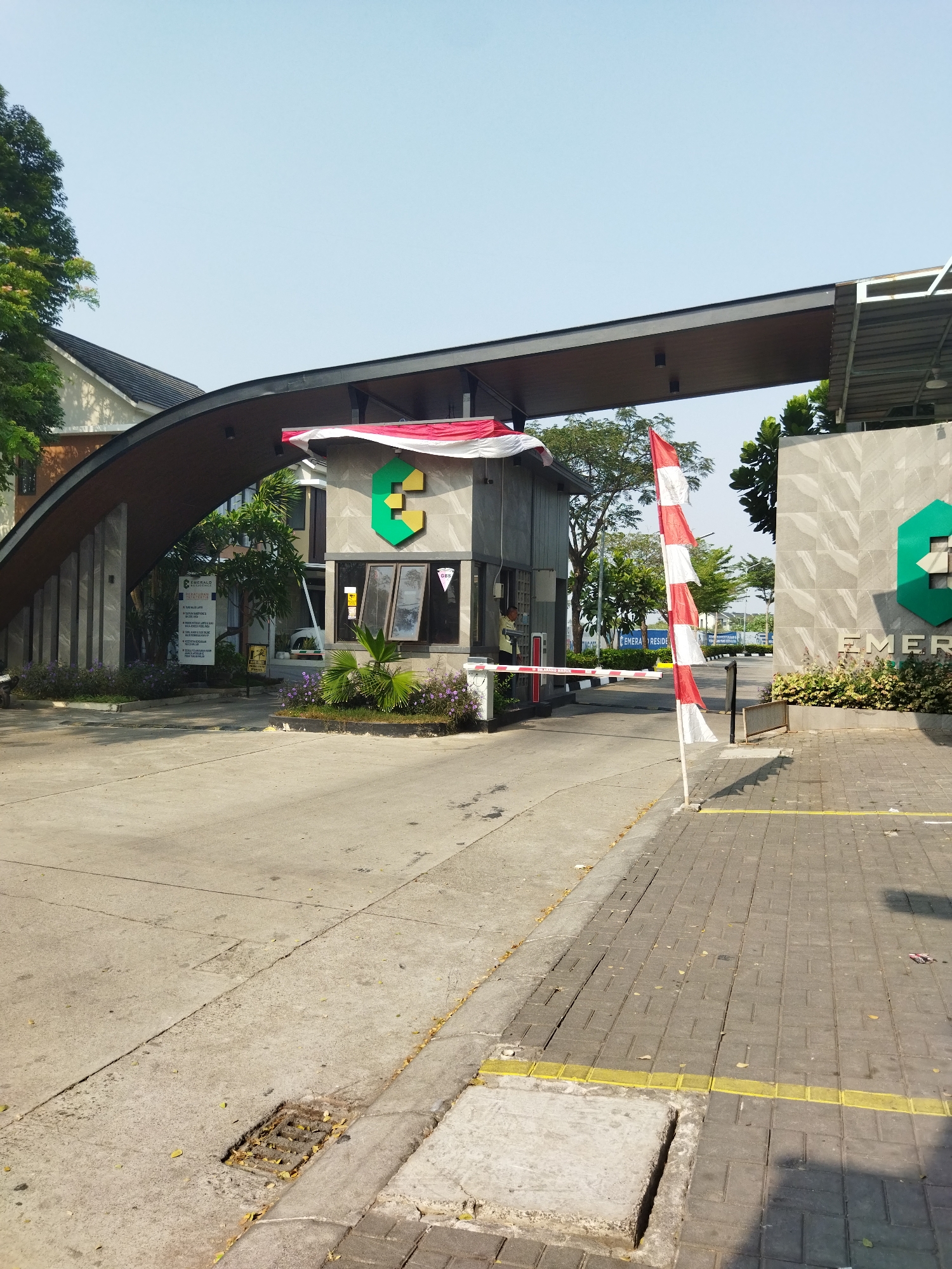 The image captures the entrance to Emerald Residence in Kosambi, Indonesia. The scene is dominated by the architectural features of the entrance gate, which includes a large, curved, dark-colored overhang supported by stone pillars on the left and a gray, stone-clad structure on the right. The structure on the right houses a security booth with windows and a barrier gate. A red and white Indonesian flag is prominently displayed on a pole to the right of the security booth, waving slightly.
A security guard, wearing a light-colored shirt, is visible inside the booth, appearing to interact with someone or operate equipment. A barrier arm, painted with red and white stripes, is lowered across the entrance road. The road itself is paved with asphalt and concrete, with a sidewalk made of interlocking pavers on the right side. Yellow tactile paving lines are visible on the sidewalk.
Vegetation is present, including trees and flowering plants, particularly around the base of the entrance structures and along the roadside. A sign is mounted on a pillar to the left of the entrance, displaying information related to Emerald Residence. In the background, more trees and buildings are visible under a clear blue sky, suggesting a well-maintained residential or commercial complex. The overall impression is one of a secure and organized entrance to a property.