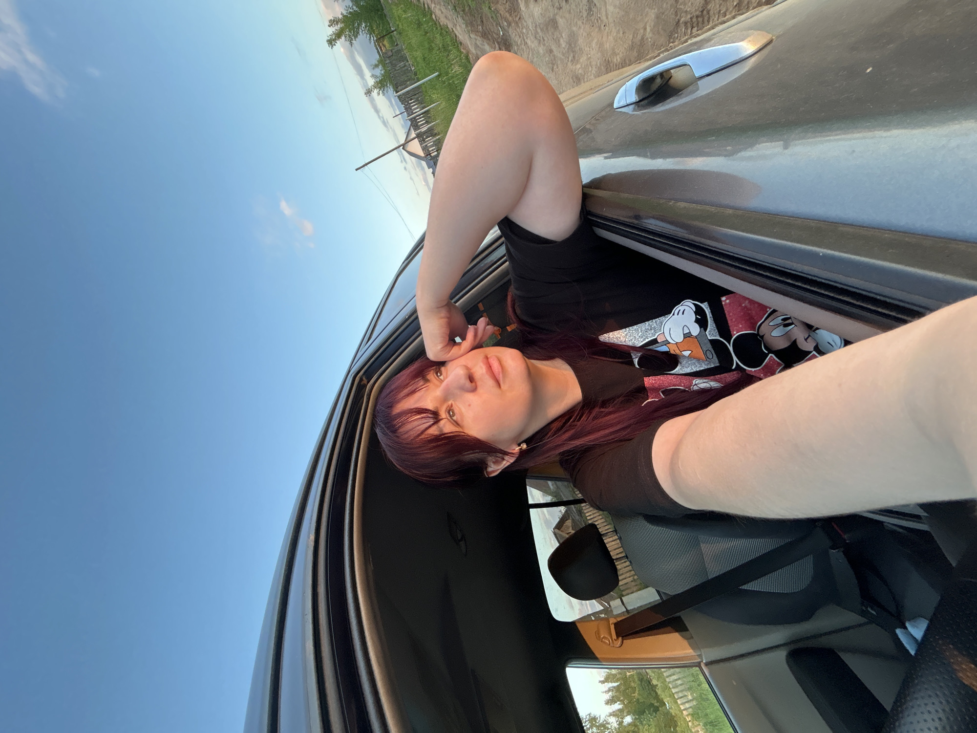 The image captures a young woman with dark hair and bangs, wearing a black t-shirt with a graphic print of Mickey Mouse. She is sitting in the passenger seat of a car, with her left arm resting on the open car door and her head tilted upwards, looking out. Her right hand is touching her temple. The car's interior is visible, including a seatbelt and the back of the driver's seat. Outside the car, the scene suggests a rural or semi-rural environment. A dirt road is visible, along with a wooden fence and some vegetation, including trees and bushes. In the background, a simple house with a roof can be seen. The sky is a clear blue with a few wispy clouds, and the lighting indicates it is late afternoon or early evening, possibly during sunset, casting a warm glow on the scene. The presence of a dirt road, simple fencing, and the overall landscape are consistent with a location in a town like Фоки, Russia, which is known for its natural surroundings and less urbanized character.