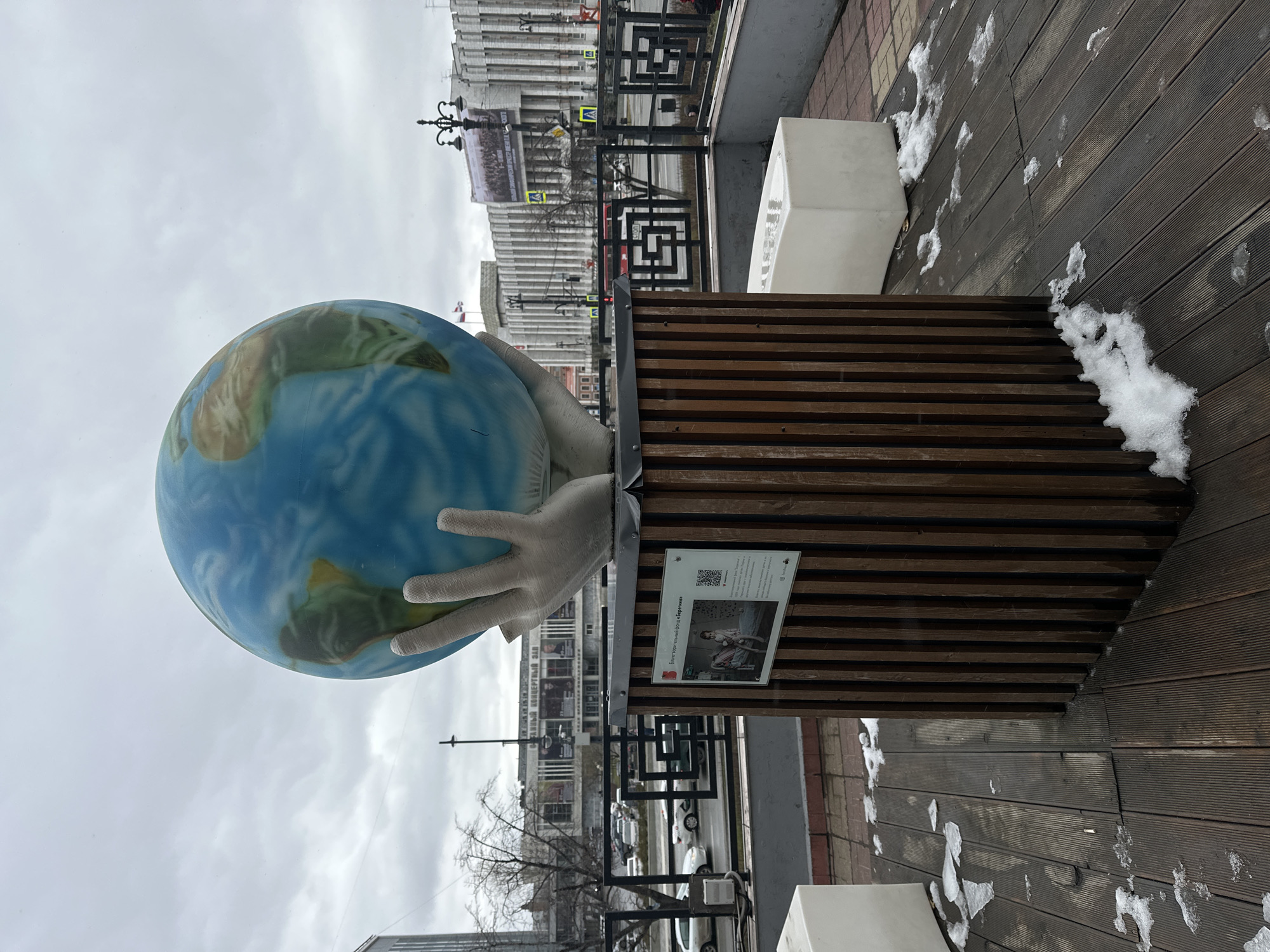 The image depicts a sculpture of two hands holding a globe, placed on a wooden structure. The structure has a plaque with images and QR codes affixed to it. The sculpture is located in an urban setting, with buildings visible in the background, including a large building with a banner across its facade. The ground is wooden decking with patches of snow on it. The sky is overcast. Based on the plaque mentioning "Благотворительный фонд Дедморозим" and the style of architecture, this image is likely taken in Perm, Russia.