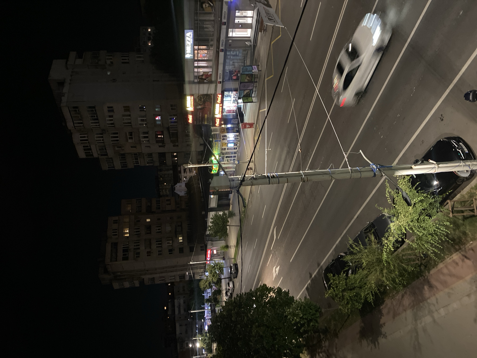 The image depicts a nighttime street scene in Iași, Romania. A multi-lane road runs through the center of the frame, with several cars visible. One white car is in motion, appearing to be driving at speed and leaving a light trail of smoke or dust. A black car is parked on the side of the road, partially obscured by trees.
On the right side of the road, there are several buildings with illuminated storefronts. Signs for "MUZZZA," "BRD," and "Coca-Cola" are visible. One building has multiple floors with windows, some of which are lit from within.
On the left side of the road, there are more buildings, including apartment blocks with illuminated windows. Trees line the sidewalk. A traffic light pole with wires is prominently positioned in the foreground. The overall impression is that of a busy urban street at night.