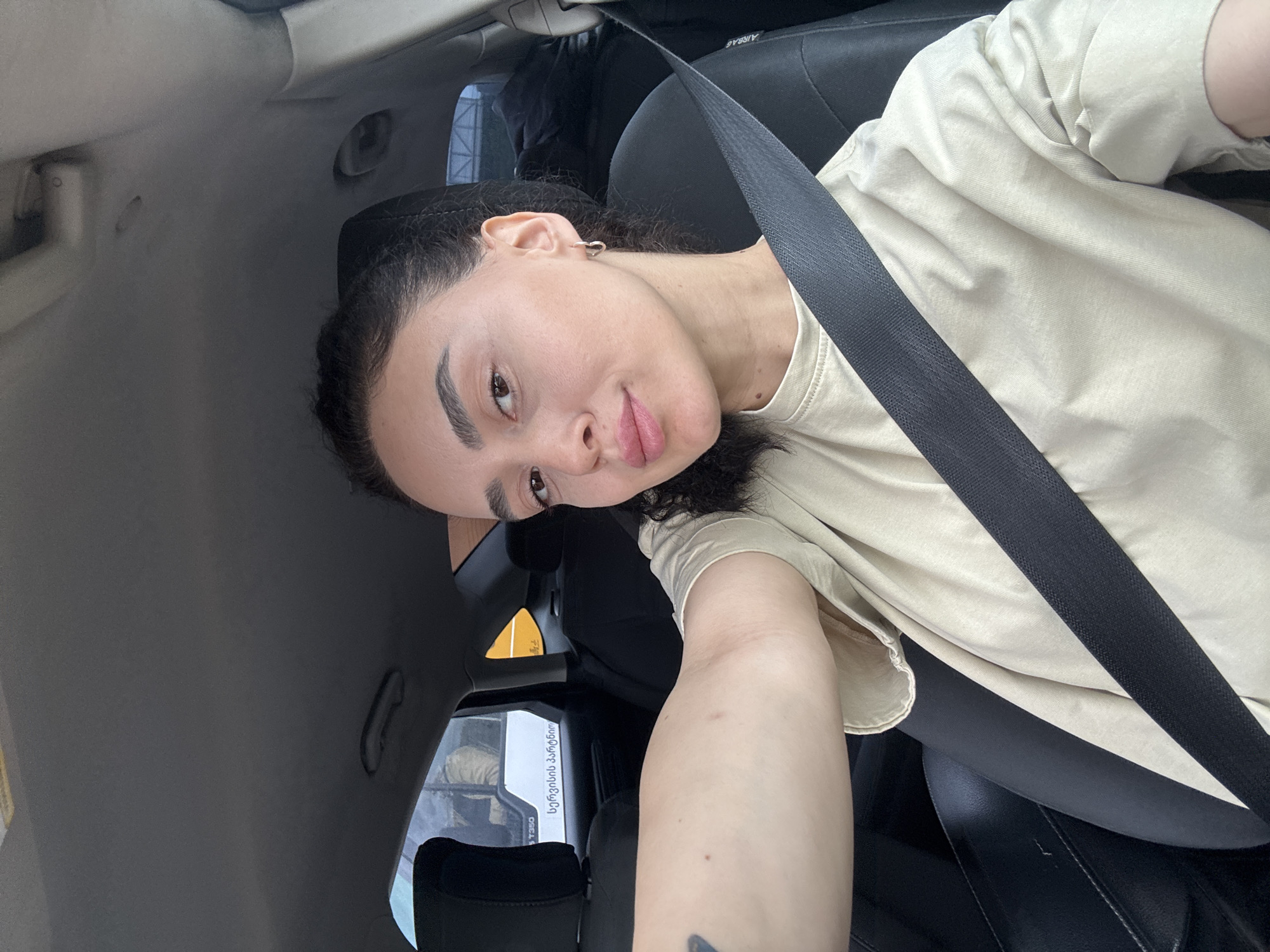A selfie from inside a car shows a young woman wearing a seatbelt and a light beige t-shirt. The car’s interior is primarily gray and black. Outside the car window, partially visible in the lower left of the frame, is a bus with Georgian script. The location is Tbilisi, Georgia, based on the language on the bus.