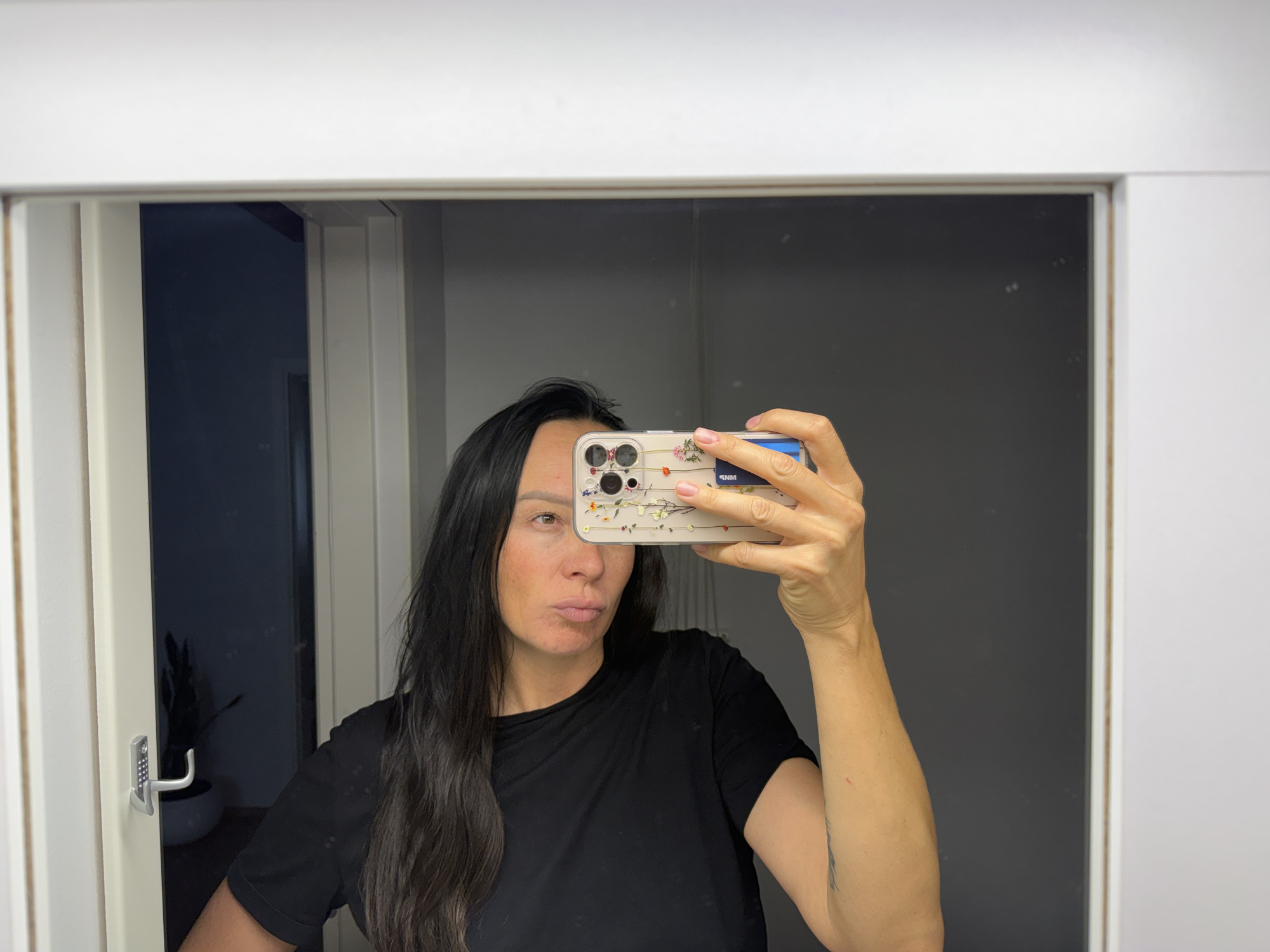In the reflection of a full-length mirror, a woman with long, dark hair and a black t-shirt is taking a selfie with her phone. The phone case has a floral pattern. She is puckering her lips. To the left of the mirror, a doorway is visible with a silver door handle and a keypad. Beyond the doorway, a dark room is visible with a plant in a pot. The mirror is framed by a white border. The background appears to be an indoor setting, possibly a hallway or dressing room. The image does not provide any specific information about the city of Chaikovsky, Russia.