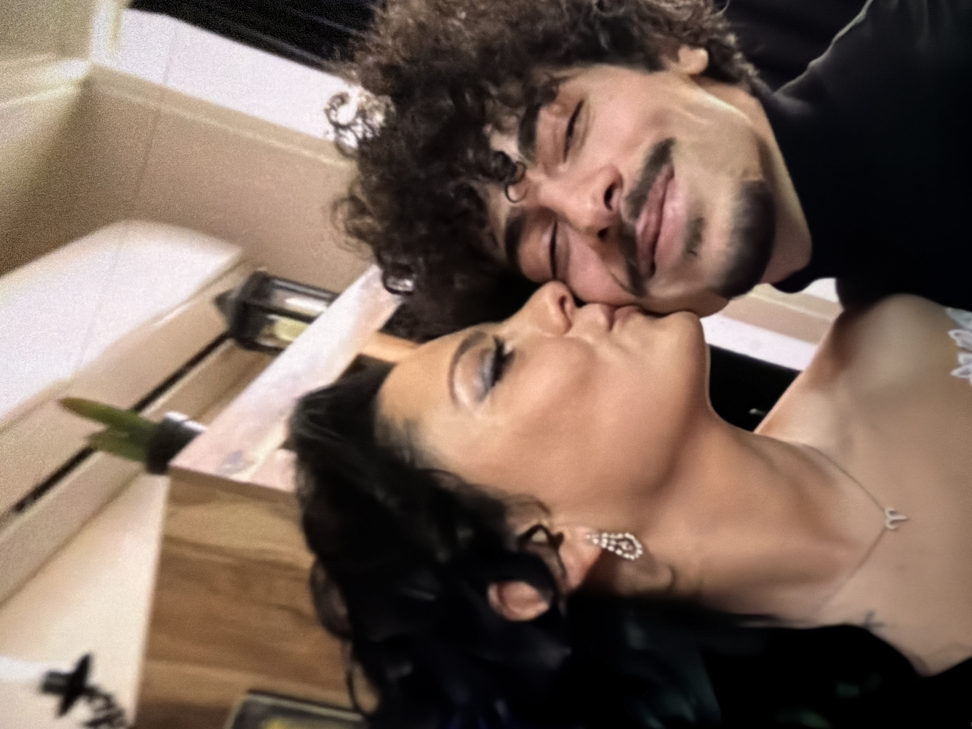 A close-up, slightly angled shot shows a man and a woman in an intimate moment. The man, with dark curly hair and a mustache, has his eyes closed and appears to be kissing the woman. The woman, with dark hair, is also leaning in. She wears dangling earrings and a delicate necklace. The background features blurred elements, possibly indicating an interior setting. The location is Unknown, Unknown.