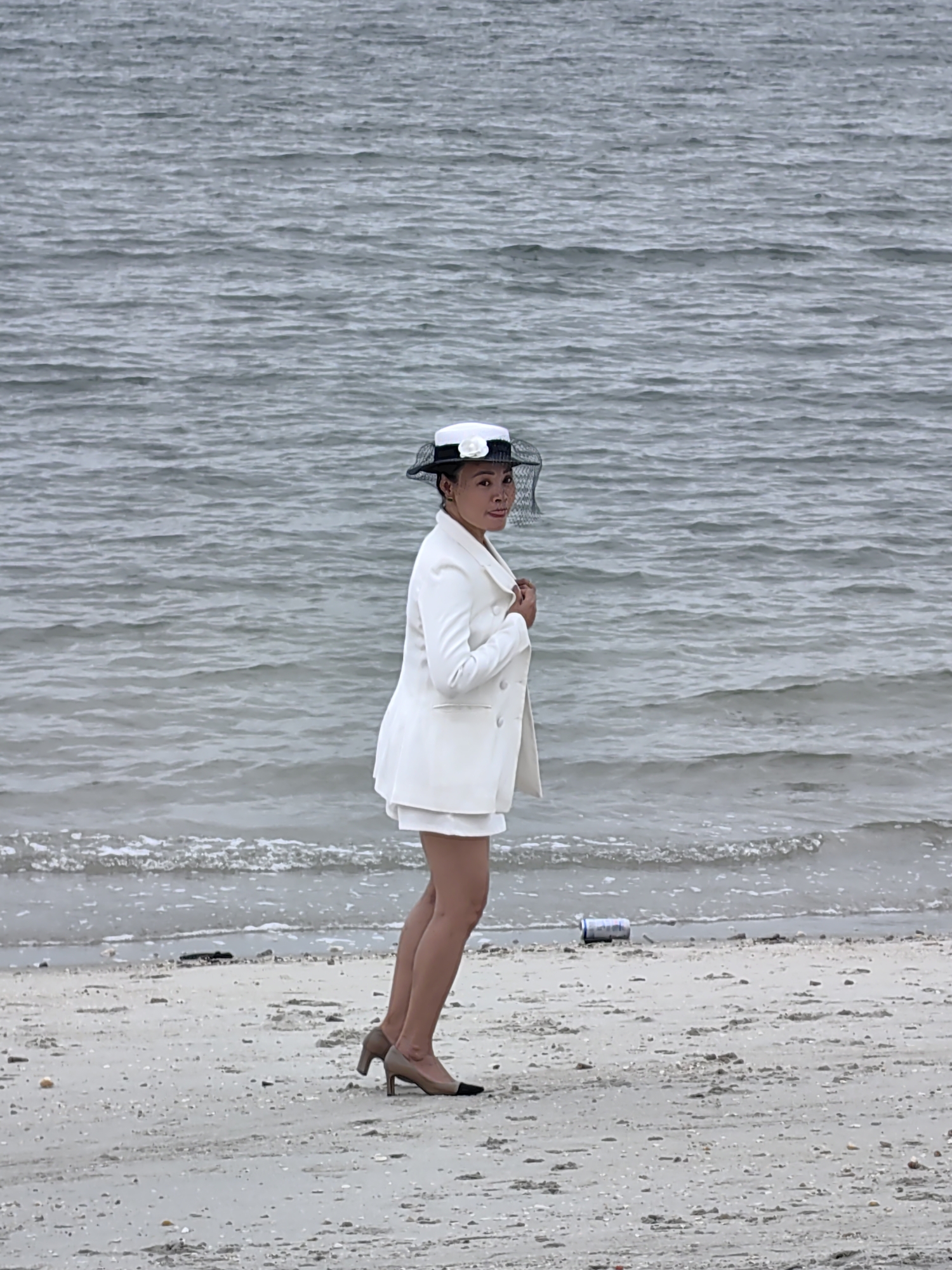 An Asian woman, appearing middle-aged, stands on a sandy beach next to the water. She is dressed in a formal-casual white ensemble, consisting of a white double-breasted blazer and matching white shorts. On her head, she wears a black and white hat with a decorative white flower and a black mesh veil partially covering her face. Her footwear consists of high-heeled, two-toned pumps, light brown/beige with black toe caps. She stands in a slight side profile, looking over her right shoulder directly at the camera with a neutral expression.
The background features a large body of water, likely a sea or bay, with gentle, gray-toned ripples stretching to the horizon under what appears to be an overcast sky. Small waves are breaking softly on the shoreline. The beach itself is composed of light-colored sand, scattered with small shells and dark debris. Near the water's edge, to the woman's right, lies a discarded metallic beverage can. The scene suggests a posed photograph in a coastal environment, specifically in Bai Chay Ward, Vietnam. No discernible text is visible on any objects.