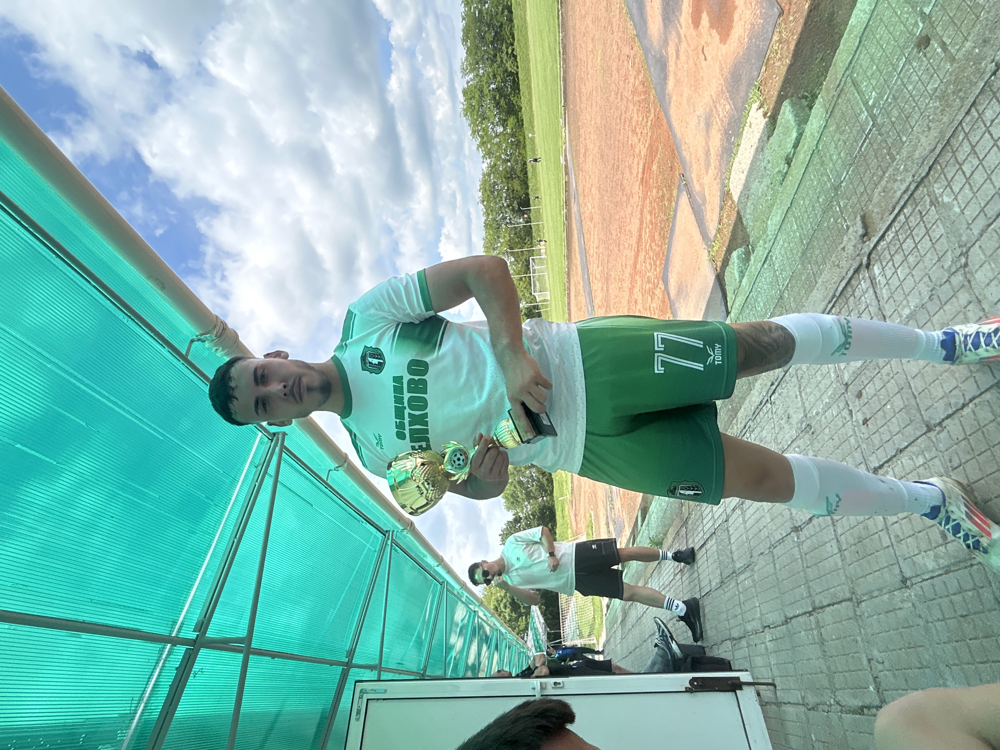 A man wearing a soccer uniform stands in a covered area, possibly a dugout, holding two trophies. He is wearing a white jersey with green accents and the number 77 on his shorts. The inscription on his jersey is in a Cyrillic alphabet, potentially indicating a Slavic or Eastern European origin. The background shows a soccer field, with another individual in a white shirt visible further back. The sky is overcast. The location details, "Unknown, Unknown" suggest the exact place is unspecified or unknown.