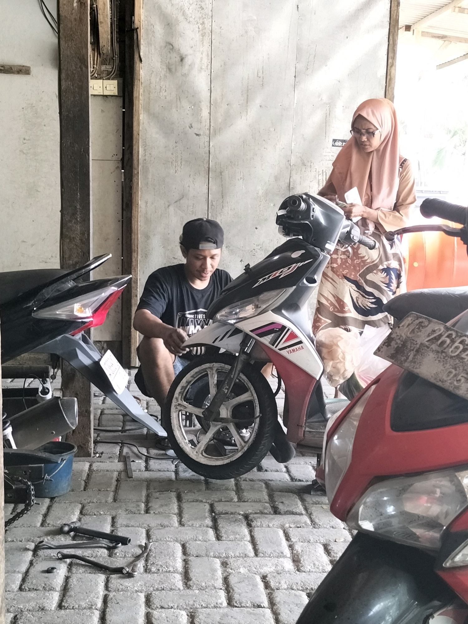 In an outdoor workshop located in Tasikmadu, Indonesia, a man in a black t-shirt and baseball cap is crouched down, appearing to work on the front fender of a white and red scooter. He is holding a tool in his right hand, focused on the scooter. Beside him, a woman wearing a light orange hijab and glasses stands, holding a white object, possibly a rag or a part, near the handlebars of the same scooter. Behind them, another scooter, predominantly red, is partially visible. The ground is paved with grey interlocking bricks, and various tools, including a hammer and some metal rods, are scattered on the ground. The background consists of a wooden structure and a light-colored wall, suggesting an open-air or semi-enclosed workspace.