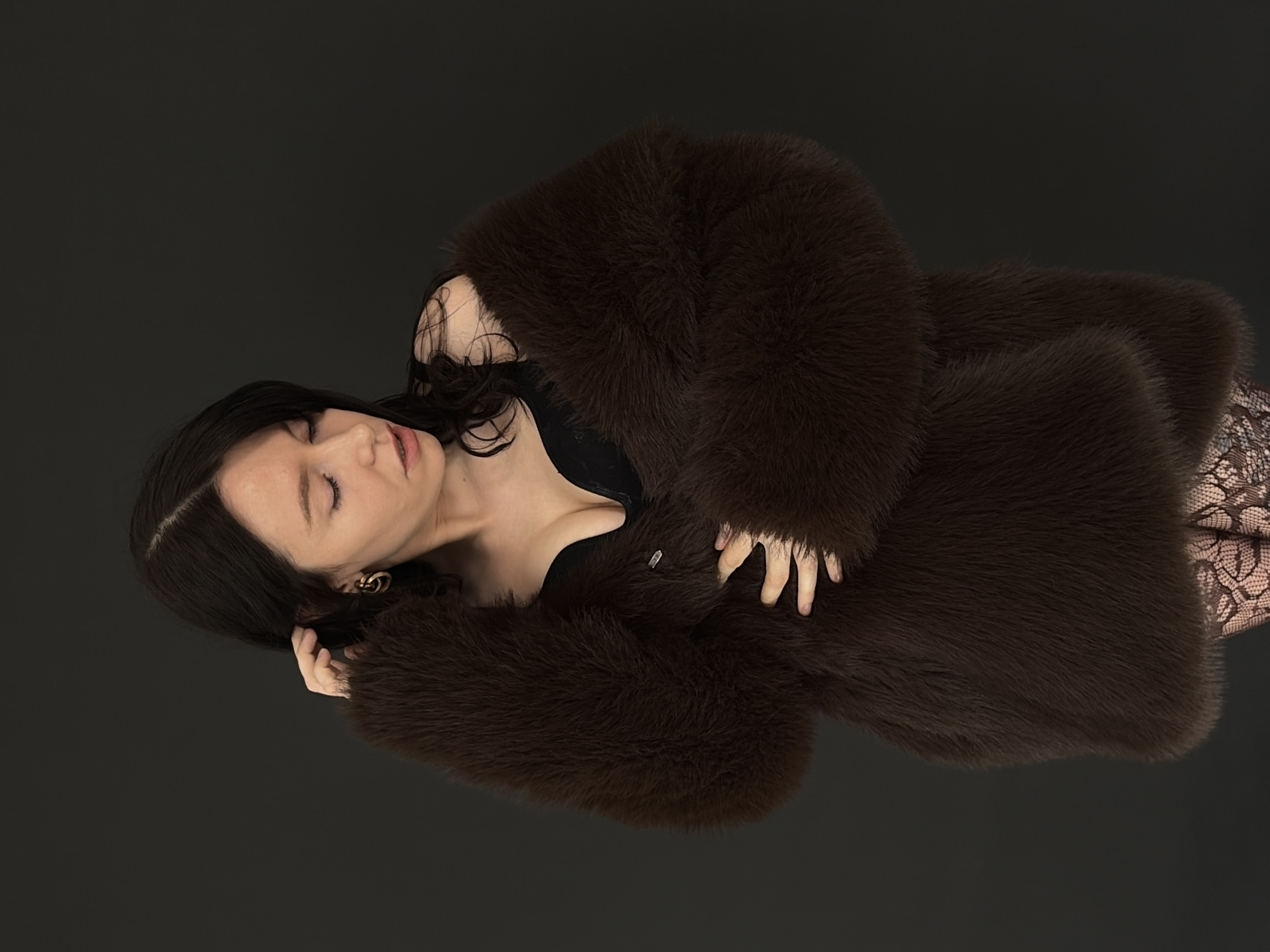 A woman with dark hair is reclining on a dark gray surface, partially covered in a dark brown fur coat. She is wearing a black top and patterned tights. Her eyes are closed, and one hand is resting on her head while the other is on the coat. A gold earring is visible in her left ear. There is no indication of the location being Chaikovsky, Russia, visible in the image.