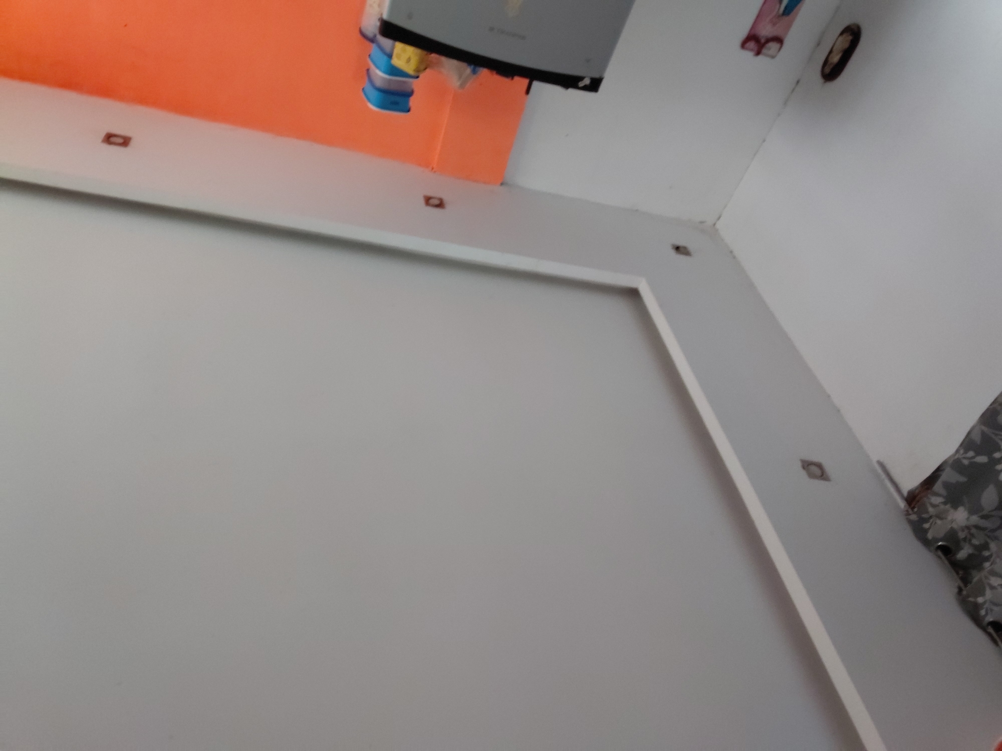The image captures an indoor scene, likely a room in a residence in Santa Ana, Venezuela, viewed from a low, tilted angle looking upwards. The dominant colors are light gray/white for the walls and ceiling, with a section of the wall or ceiling painted bright orange.
A white, rectangular trim or border creates a frame-like element on the light gray wall, possibly outlining an area for a television or a decorative panel. Along this trim and on the ceiling, several small, dark, square-shaped fixtures are visible, some with a central circular element, suggesting recessed spotlights or decorative accents.
Attached to the orange-painted section of the wall/ceiling is a gray electronic device, possibly an inverter or an air conditioning unit. Below this device, bundled blue, yellow, and clear plastic or rubber components are visible, likely related to its installation or wiring. Faint text is visible on the gray device, possibly a brand name, but it is not clearly legible.
In the upper right corner, an oval-shaped dark opening is present on the white ceiling, potentially for a light fixture or ventilation. Further right, a piece of pink or red material with a visible letter "B" is attached to the wall. In the bottom right corner, a portion of a gray fabric with a white floral or leaf pattern, likely a curtain, is visible.
No people are present, and no specific activity is taking place. The lighting appears consistent with an indoor setting, possibly with artificial light or indirect daylight.