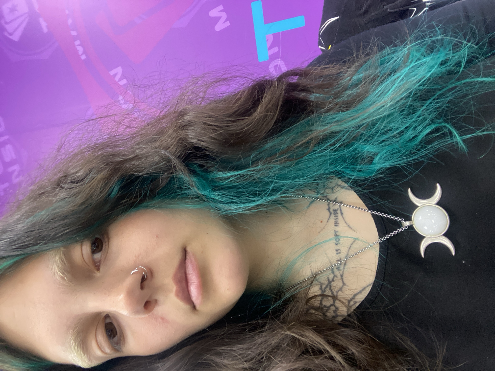 A young woman is visible with brown and teal hair. She has fair skin and brown eyes, with a ring piercing in her nose. Around her neck, she wears a silver chain with a white moon pendant, with a visible tattoo on her neck. A background in shades of purple has printed text, and a blue 'T' shape is visible on the right.