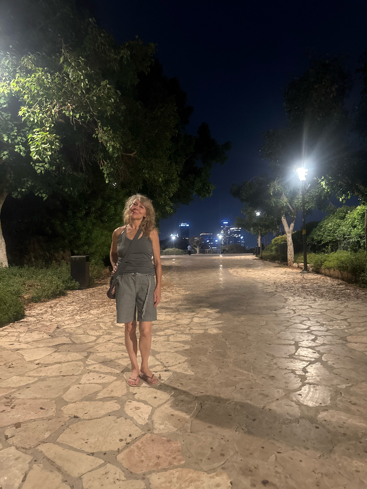 A full-body, eye-level shot shows a woman standing on a stone-paved walkway at night in Gan HaPisga, Ramat Gan, Israel. The woman is facing the camera with a slight smile and looking upwards. She has long, blonde, wavy hair and is wearing a grey tank top and grey shorts. She is also wearing pink flip-flops and has a dark-colored purse slung across her body. The walkway is illuminated by streetlights and the light from the city skyline in the distance, which is visible behind her. There are trees and foliage on either side of the walkway. The city skyline features numerous illuminated buildings. The overall mood of the image is calm and serene.