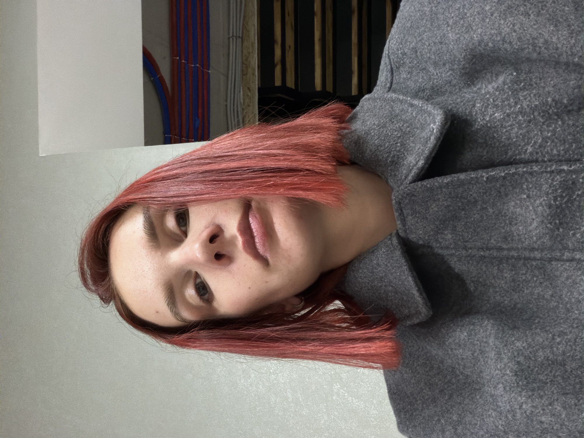 A close-up, rotated portrait shot of a person with dyed red hair wearing a gray woolen coat. The background is a combination of a light gray wall on the left, with electrical conduit on the top left and some dark brown or black vertical elements behind the person on the right. There is no way to ascertain the city of Chaikovsky, Russia, from this image.