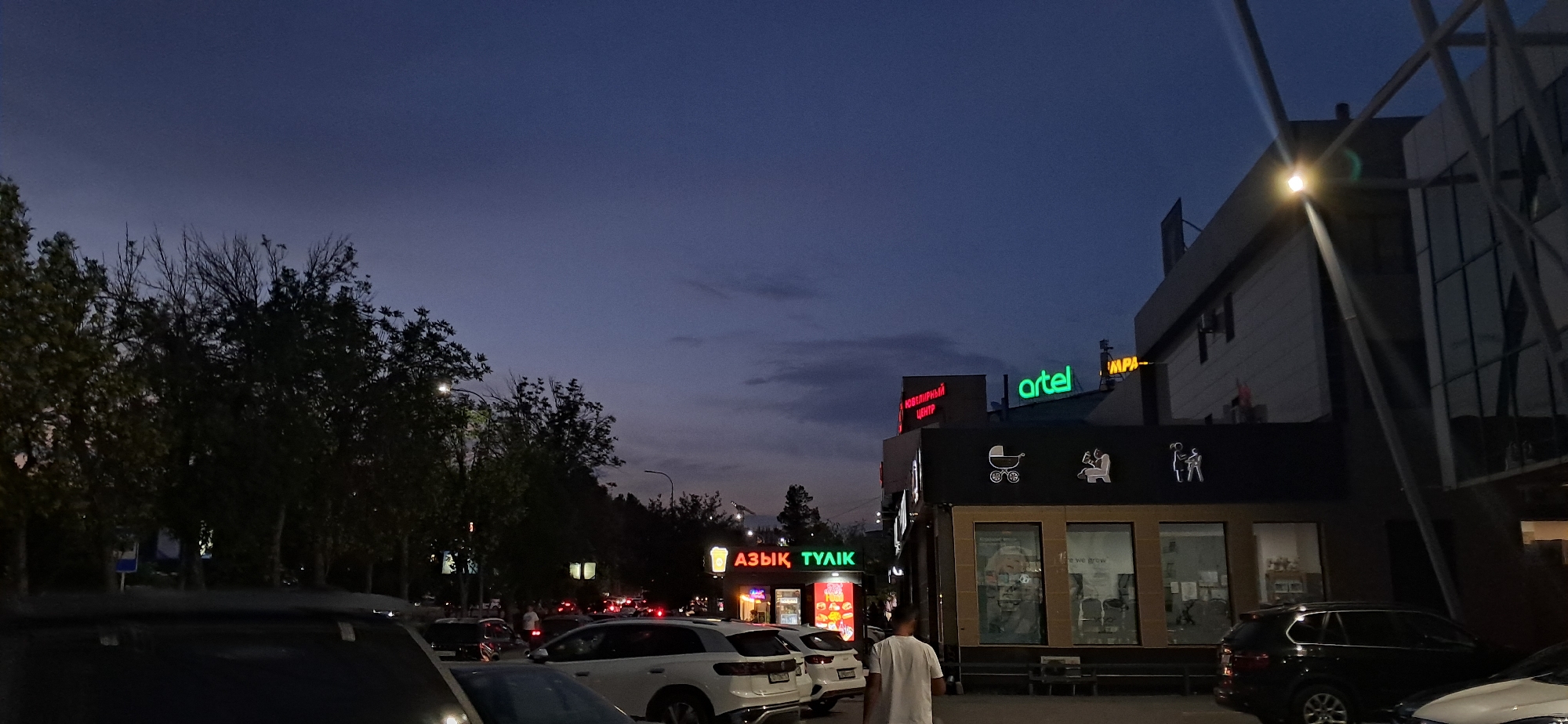 The media file captures an outdoor scene at dusk. The sky is a deep indigo, with hints of purple and scattered clouds, suggesting the transition from day to night. In the foreground, several cars are parked, with the hood of one vehicle prominent on the left. Behind the cars, trees line the street, their branches silhouetted against the twilight sky.
Several commercial buildings are visible, adorned with neon signs. One sign displays the word "artel" in bright green letters. Another sign, in red neon, reads "ЮВЕЛИРНЫЙ ЦЕНТР", which translates to "Jewelry Center". To the left, another neon sign in yellow and green says "АЗЫҚ ТУАЙҚ". The buildings have large glass windows, some reflecting the fading light. On one of the buildings, there are graphic icons depicting a baby stroller and figures of people.
A few individuals are present in the scene. One person, wearing a white t-shirt, is walking away from the camera towards the buildings. Other figures are faintly visible near the parked cars and in the distance. The overall atmosphere is calm and transitional, characteristic of early evening. The lighting suggests artificial illumination from streetlights and the neon signs, competing with the diminishing natural light.