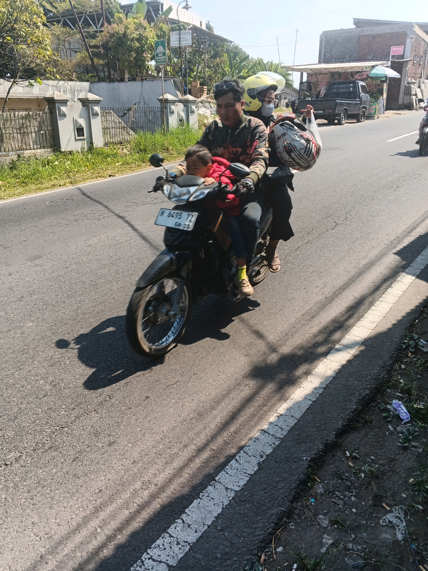 Three individuals are riding on a motorcycle on a paved road in Bandungan, Indonesia. The person in the front is a child wearing a red jacket. Behind the child is an adult male wearing a camouflage jacket and a yellow helmet. The third individual, a man wearing a grey jacket and a white and red helmet, is seated behind the driver. The motorcycle has a license plate displaying "H 6495 TZ". The environment shows a roadside with grass, buildings, and trees. The sun is shining, casting shadows on the road.