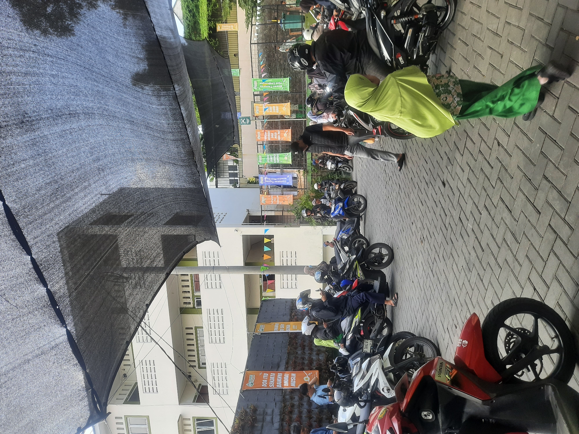 The image depicts an outdoor scene in Cimenyan, Indonesia, featuring a paved area filled with parked and some occupied motorcycles. A dark, mesh-like fabric canopy stretches across the upper portion of the frame, casting shadows. In the background, a white multi-story building with green accents and numerous rectangular windows is visible. Along a wall behind the motorcycles, several colorful banners with text are displayed. A group of people are present, some seated on motorcycles and others standing or walking. Many are wearing helmets, indicating they are likely about to depart or have just arrived. One individual in the foreground is wearing a bright green hijab and patterned skirt. The overall atmosphere appears to be one of a gathering or waiting area.