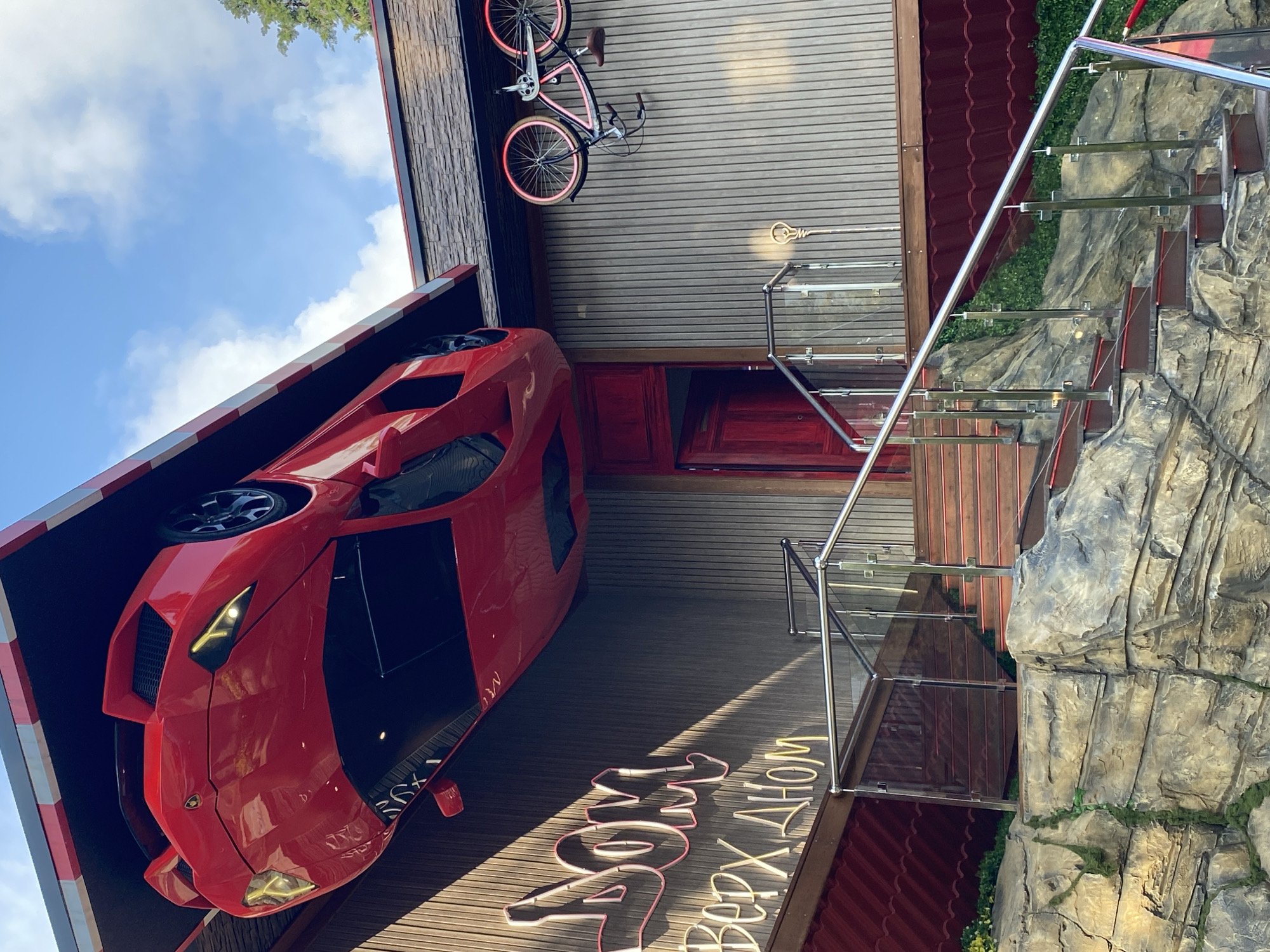 The image depicts an upside-down house attraction. A red car, presumably a display model, is attached to the roof, along with a bicycle. A set of stairs with glass railings leads to a red door. The building features a simulated rock façade. The name of the attraction, "ДОМ ВВЕРХ ДНОМ," in Russian, is written in neon lettering on the floor. The location is in the city of Sochi, Russia.