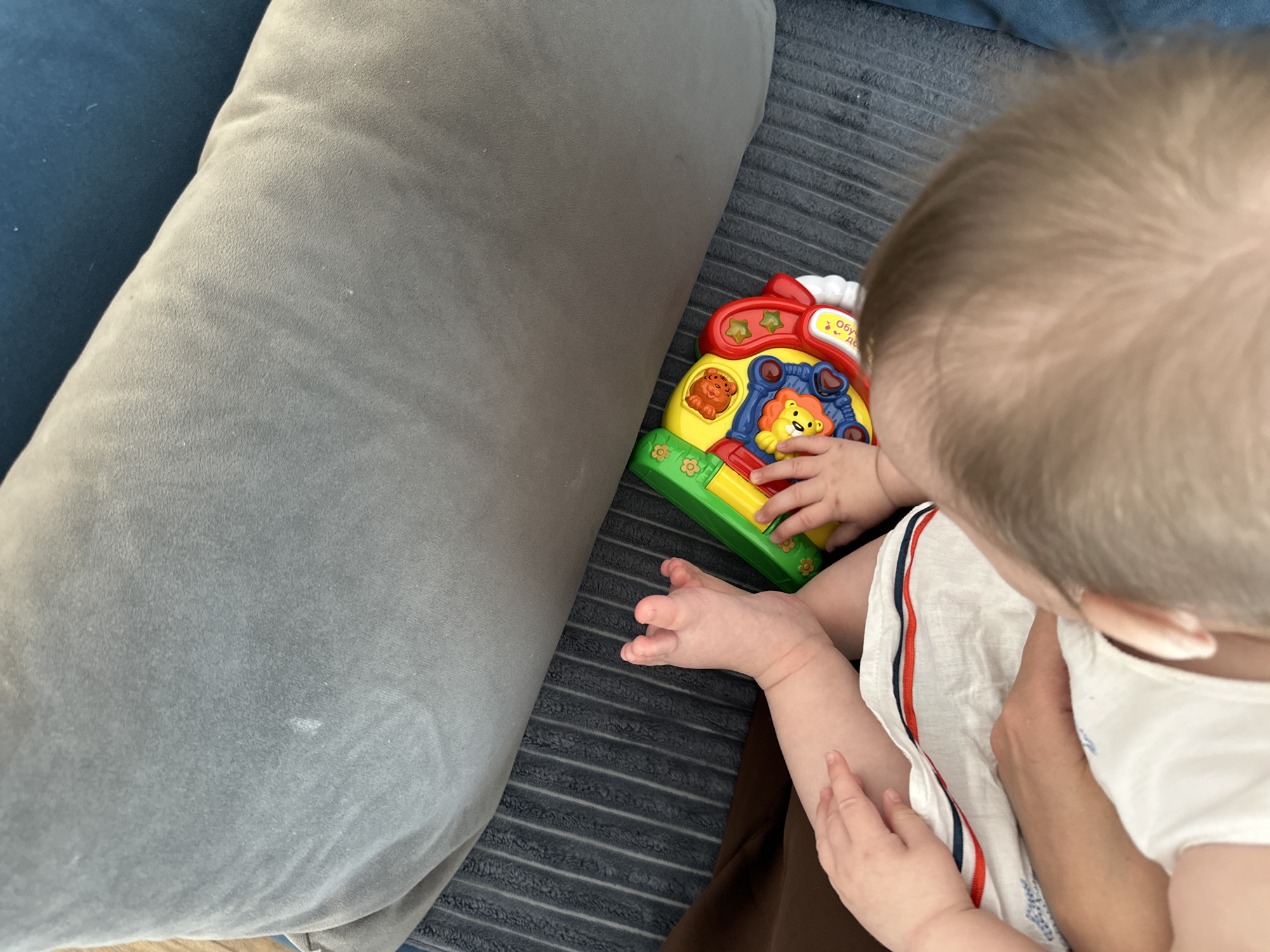 A child is seated on a striped gray surface next to a gray cushion or pillow. The child is wearing a white garment with red and blue stripes. The child is holding a colorful toy with animal illustrations. The setting appears to be indoors. There is no information to suggest that this image was taken in Sepych, Russia.