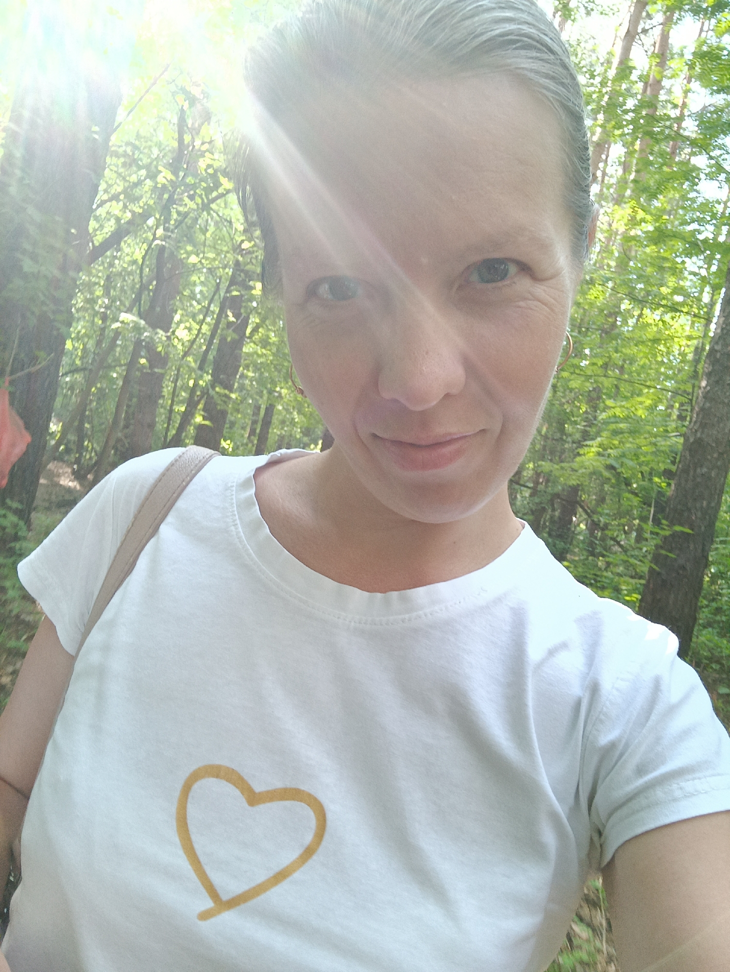 The image is a close-up selfie of a middle-aged woman in a wooded area. She is wearing a white t-shirt with a simple gold-colored heart outline on the front. She has short, graying hair and is wearing small hoop earrings. Sunlight filters through the trees in the background, creating bright highlights. The location is possibly the city of Chaikovsky, Russia, though no specific landmarks confirm it. The environment is a forest with tall trees and green foliage. There are no visible interactions or other subjects.