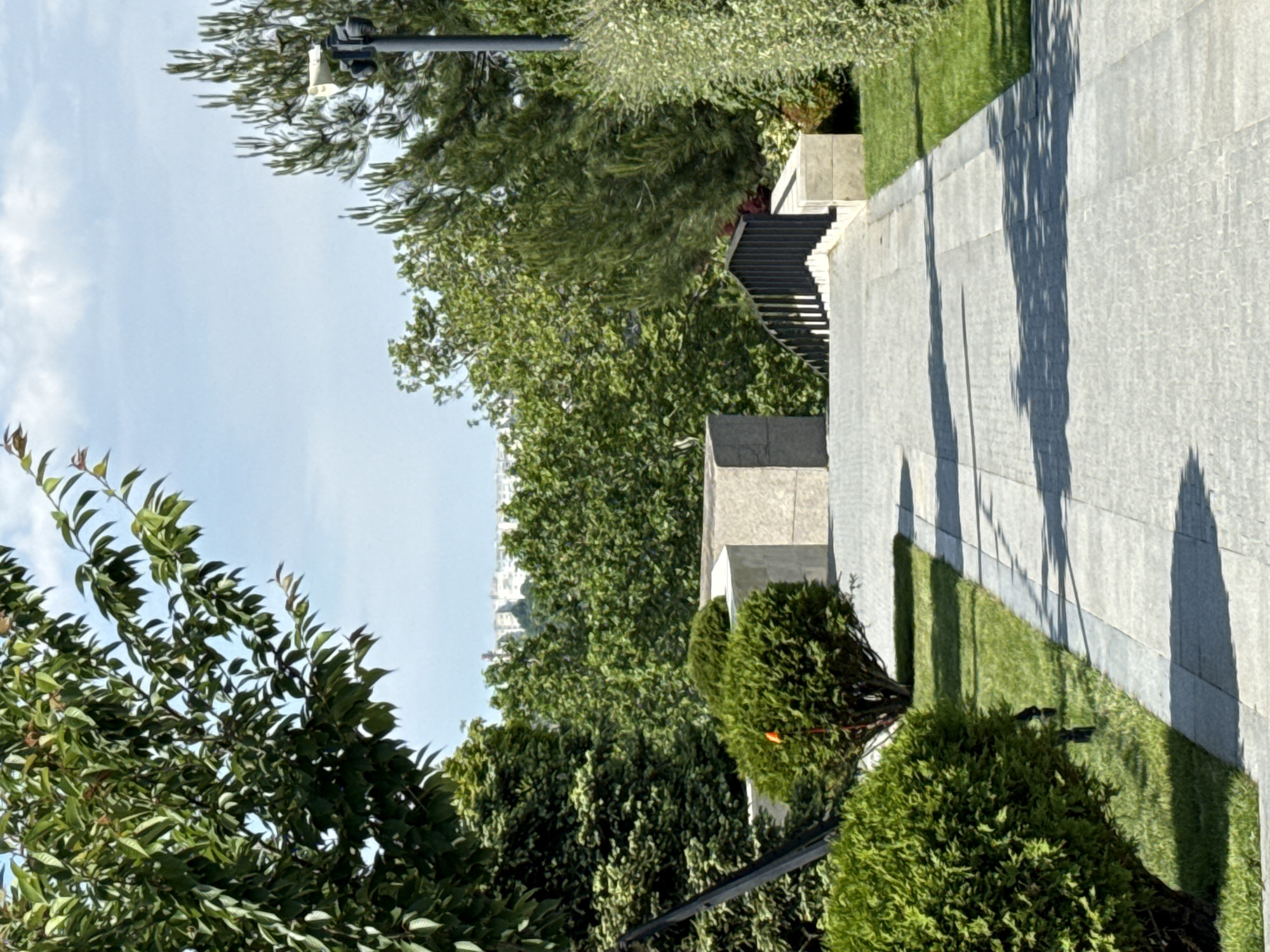 The image depicts an outdoor pathway with meticulously manicured greenery. The path, paved with light-colored stone tiles, curves gently into the distance. To the left of the path, several bushes are neatly trimmed, and a large tree with dense green foliage dominates the foreground. To the right, more greenery and a line of trees frame the scene, with a tall pole featuring a camera mounted on top visible in the background. A set of stairs with a black metal railing is situated further down the path, suggesting an elevated area or a change in terrain. The sky above is a clear blue with sparse white clouds, and a distant cityscape can be seen through the trees. The overall impression is one of a well-maintained park or garden area within an urban setting.