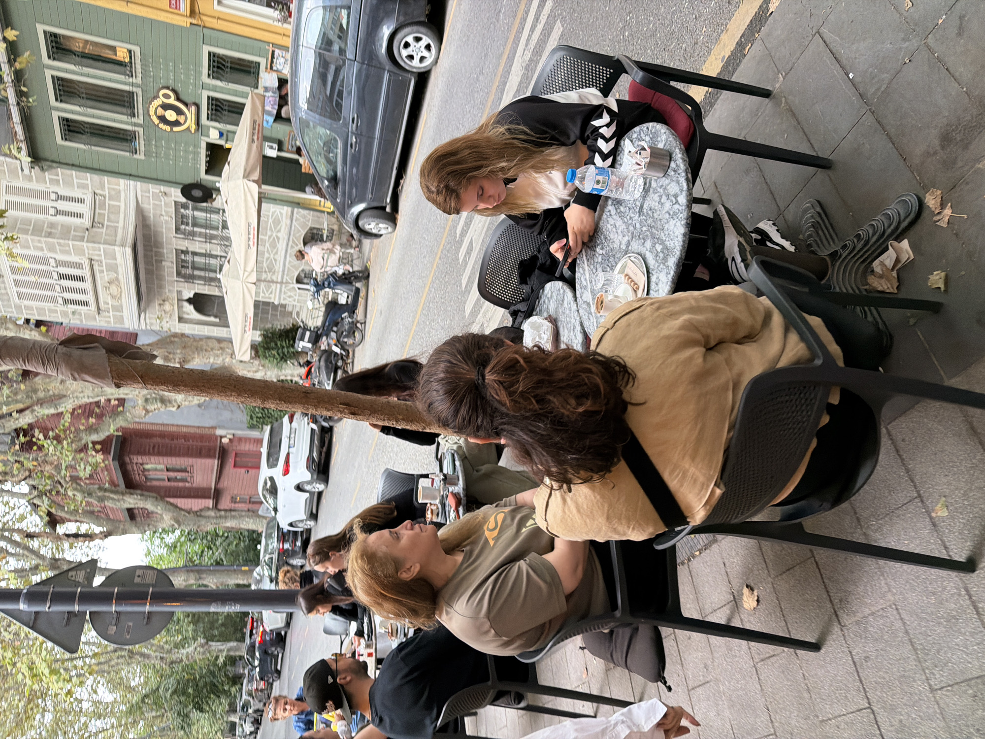 In Üsküdar, Turkey, on a paved sidewalk lined with trees and buildings, a group of people are seated at outdoor cafe tables. In the foreground, a woman with long, dark, curly hair pulled back in a bun sits facing away from the camera, wearing a tan top. To her left, a woman with blonde hair sits facing the camera, wearing a black and white jacket and looking at her phone. A bottle of water and a cup of coffee are on the marble-topped table in front of her. To the left of the blonde woman, a woman with wavy blonde hair and a tan t-shirt is seated and looking to her right. Further to the left, a man wearing a black t-shirt and a baseball cap is seated. In the background, cars are parked along the street, and a dark gray van is driving by. The buildings appear to be a mix of historic and modern architecture, with a green building featuring a prominent sign that reads "Off The Record." The scene is set during the daytime with overcast lighting.