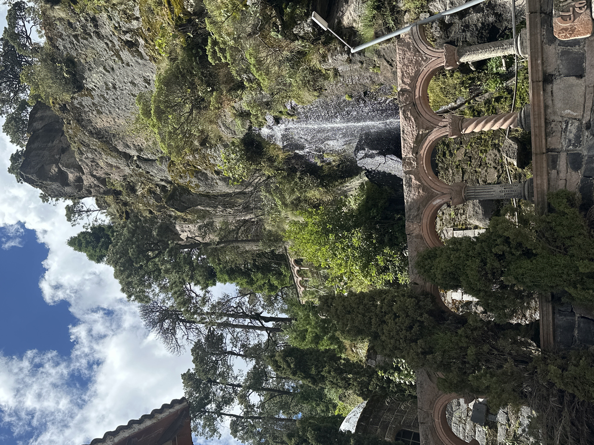 The image shows a scenic view of a waterfall cascading down a rocky cliff face, surrounded by lush greenery and tall trees. In the foreground, there is a stone archway with ornate columns, hinting at historical architecture. The sky above is a vibrant blue, dotted with fluffy white clouds. The overall impression is one of natural beauty and historical significance.
This scene is located in the city of San Mateo Ozolco, Mexico, a place renowned for its natural attractions and rich cultural heritage. The city's mountainous terrain and temperate climate make it an ideal setting for such picturesque landscapes.
