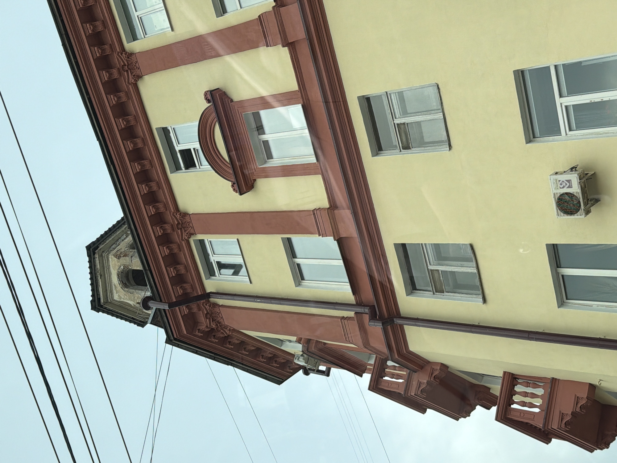 A low-angle shot depicts a multi-story building facade in Unknown, Unknown. The building features a pale yellow exterior wall adorned with red-brown architectural elements, including window frames and ornate decorations. Rectangular windows are interspersed across the facade, some with reflective glass. An air conditioning unit is mounted on the wall near a window. The top of the building includes a decorative tower-like structure. Electrical wires are visible in the foreground against a light, overcast sky.