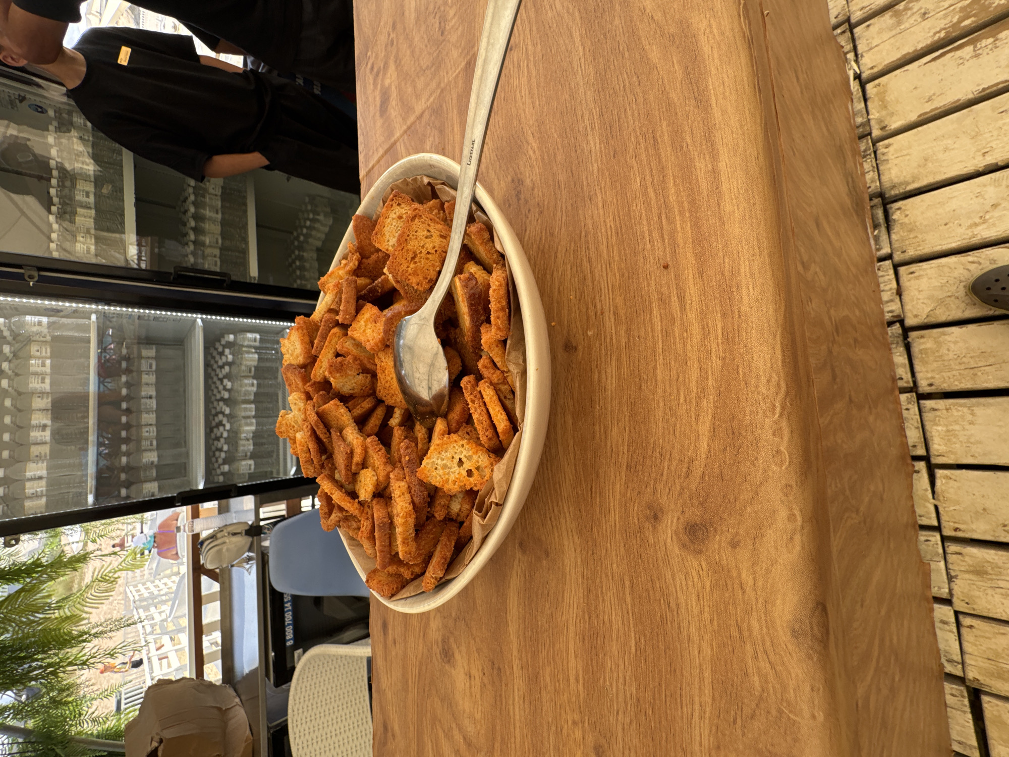 A close-up, eye-level shot shows a white bowl filled with crunchy, golden-brown croutons on a wooden table. A silver spoon rests on top of the croutons. In the background, a glass refrigerator is stocked with bottled water. Through the glass, a beach scene is visible, with people relaxing on lounge chairs and green foliage framing the top of the image. The setting appears to be a beachside establishment in Vityazevo, Russia.