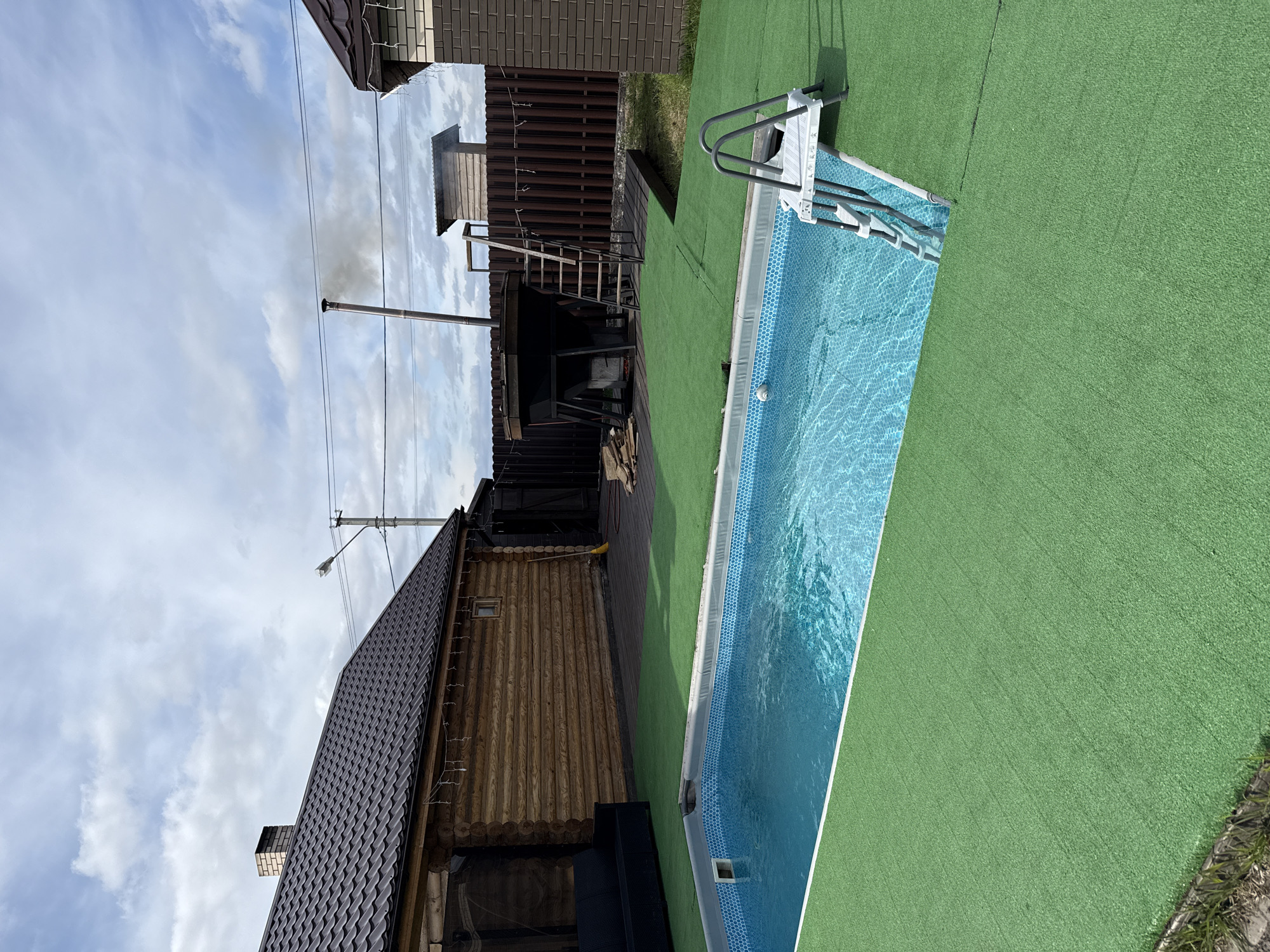 This image, taken in Солонцы, Russia, presents a backyard scene featuring a rectangular in-ground pool filled with water, surrounded by artificial green turf. A ladder provides access to the pool. Adjacent to the pool is a wooden structure with a dark roof, potentially a bathhouse or outbuilding, constructed from logs. A smoking chimney protrudes from the structure. Next to the bathhouse is an external staircase and a rusty metal fence with open shed. The sky above is partially cloudy. The image orientation is tilted.