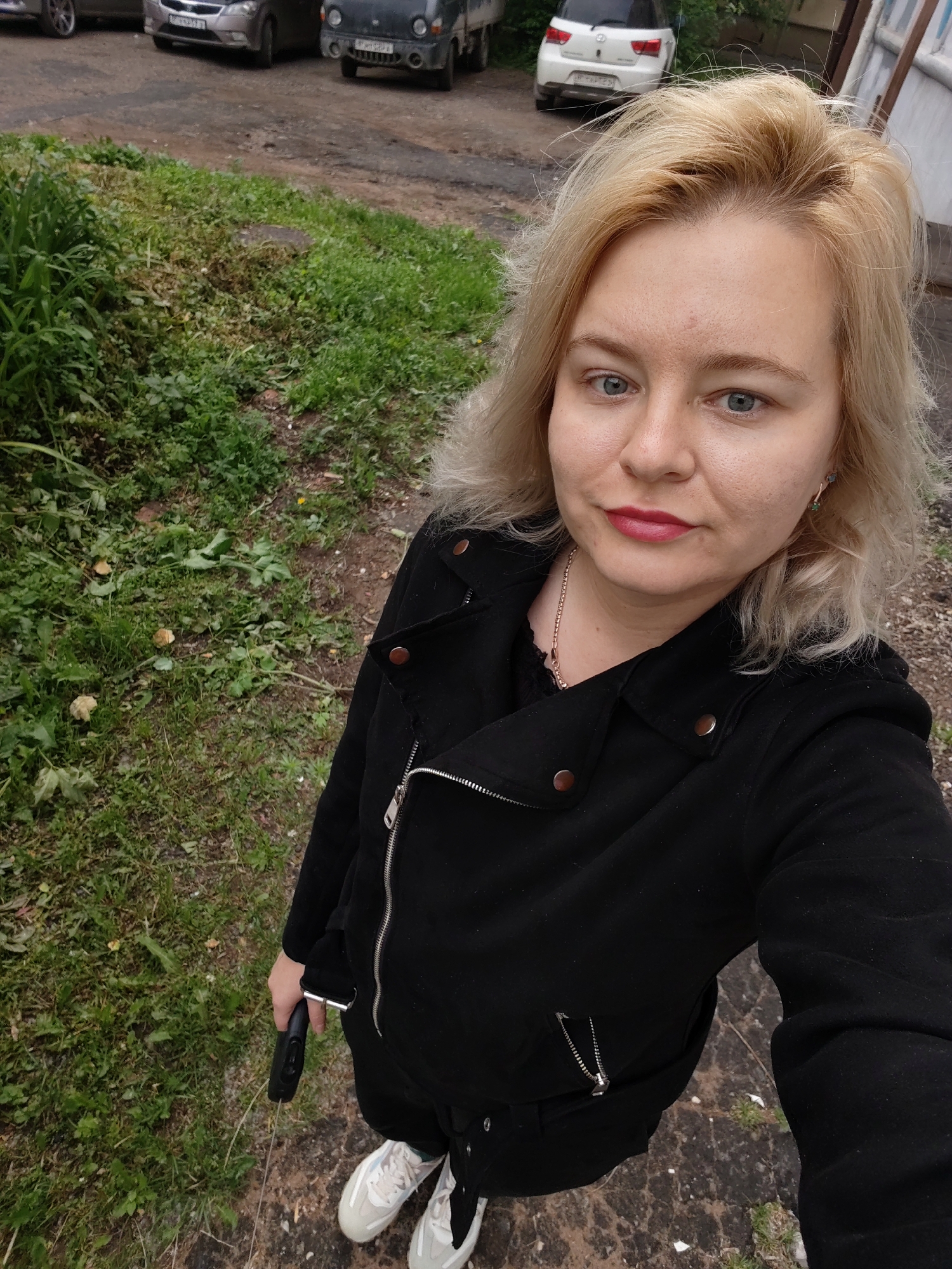 An adult female with blonde hair, blue eyes, and light complexion is depicted in a selfie-style shot, looking directly at the camera. She has red lipstick applied and is wearing a black zippered jacket with metal snaps on the lapels, a black garment underneath, black trousers, and white and grey athletic shoes. Her right hand holds a black retractable dog leash.
The scene is outdoors, set on an uneven ground composed of dirt, gravel, and patchy asphalt. A strip of unkempt grass and weeds separates the foreground from a dirt track where several vehicles are parked. In the background, from left to right, are: a partial view of a dark vehicle's front wheel and fender, a dark grey sedan with Russian license plate "B829AP 18", a white SUV with Russian license plate "A130YE 18", and a partially visible dark-colored small utility vehicle. A light-colored building wall is visible on the far right. The presence of the "18" region code on the Russian license plates identifies the location as the Udmurt Republic, confirming the scene is in the city of Izhevsk, Russia. The lighting is overcast.