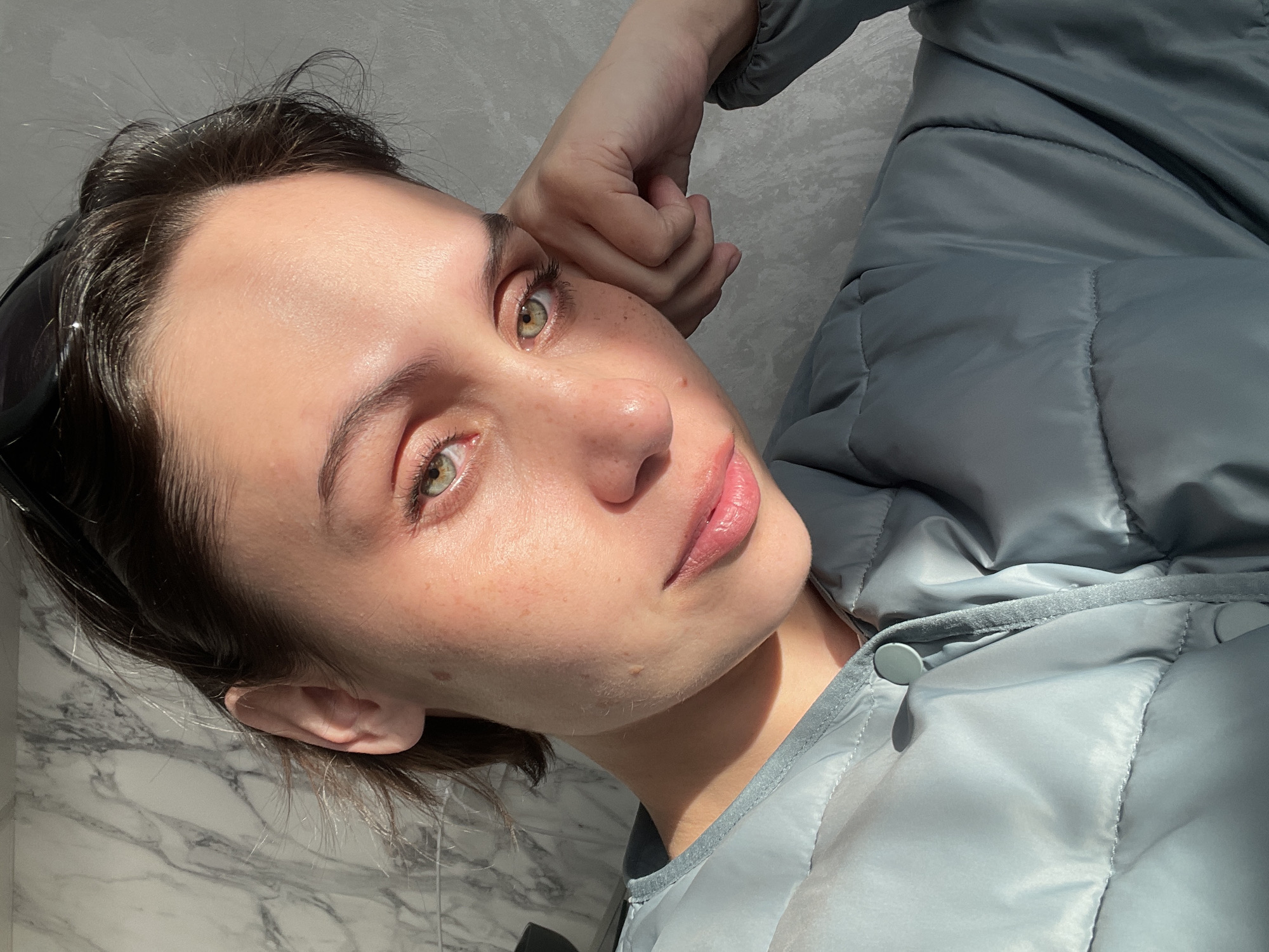A close-up, slightly canted selfie shows a young woman with fair skin, light-green eyes, and short, dark hair. She is wearing a light blue, puffy jacket. Her left hand is near her face. The background appears to be a light-colored, marbled surface, and a gray wall. There is no indication of the city or country.