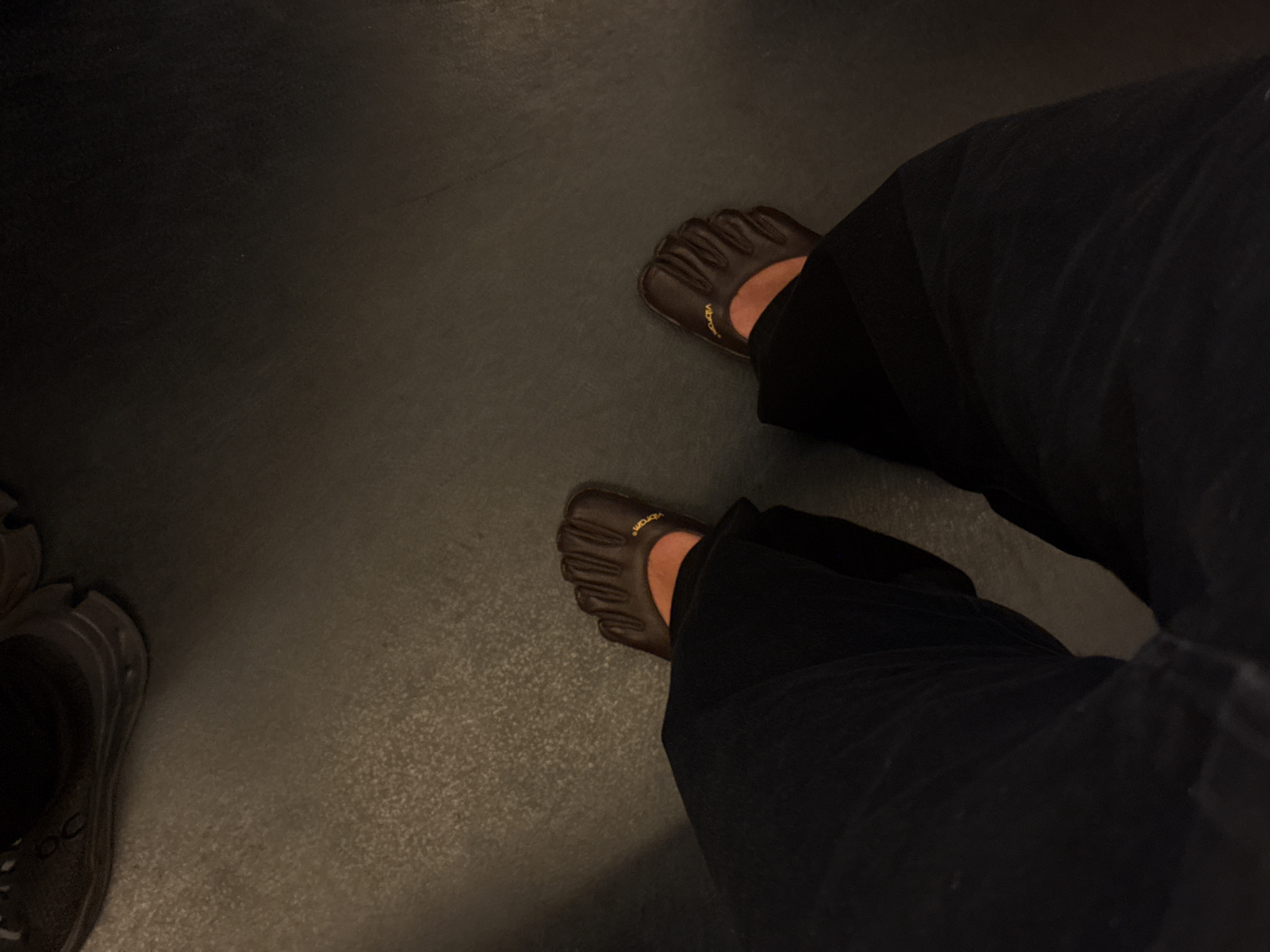 The image shows a person from the torso down, wearing dark-colored wide-legged pants and brown Vibram FiveFingers shoes. The person is standing on what appears to be a dark gray or black floor. To the left of the person's left foot, a partially visible dark gray sneaker is present. The floor appears to be clean and smooth. The lighting is somewhat dim. There is no indication of a specific location in Paris, France.