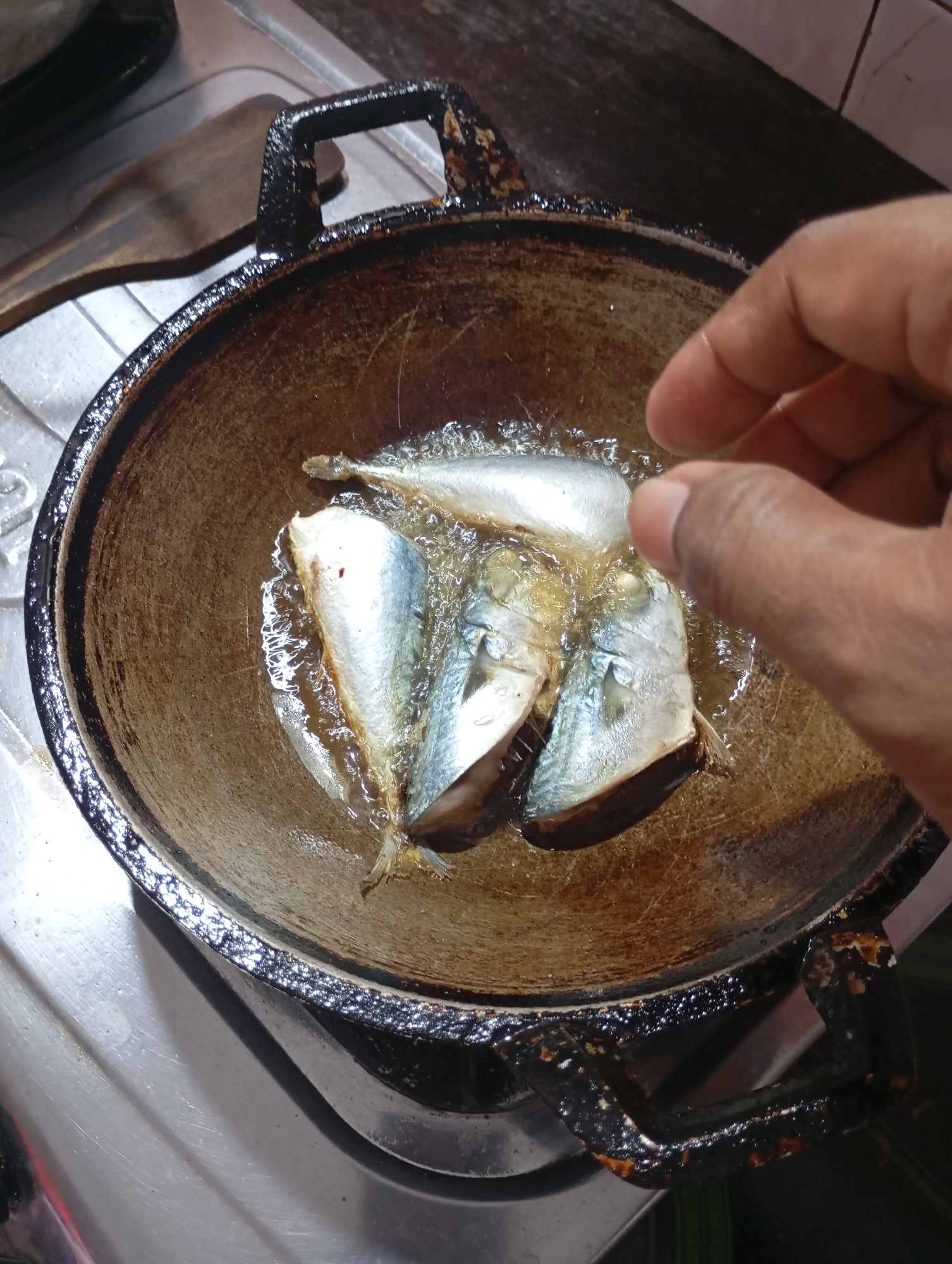 A person's hand is shown reaching into a well-used, black cast iron frying pan containing three pieces of fish sizzling in hot oil. The pan sits on a metal stovetop. A wooden spatula is visible to the left of the pan. The scene appears to be in a kitchen in Metro, Indonesia, as indicated by the context provided. The fish are light in color with silver scales and are partially submerged in bubbling oil. The hand is positioned to manipulate the fish. The pan is dark and shows signs of repeated use, with some oil residue visible on the rim and handles.